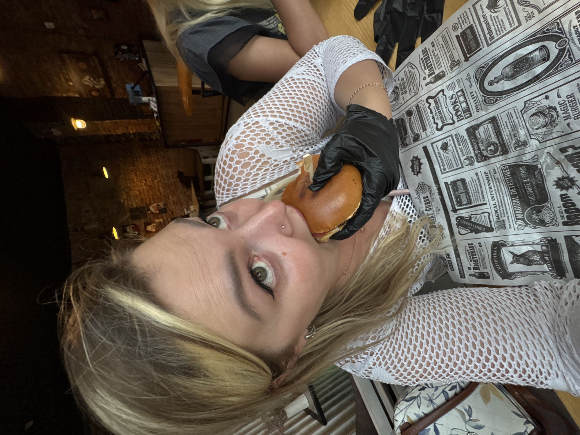 The image shows a woman in a white, crocheted top, wearing black gloves and eating a burger. She has blonde hair and is looking up and to the right. She has green eyes and winged eyeliner. She is wearing a silver earring in her left ear and a small nose stud. In the background, there is a brick wall, a counter, and other patrons. The table is covered with a newspaper-like placemat with various themed advertisements and articles, including "Nightshade" and "Magic Burger." The restaurant appears to be dimly lit with warm lighting. The overall atmosphere suggests a casual dining experience with a thematic twist.
