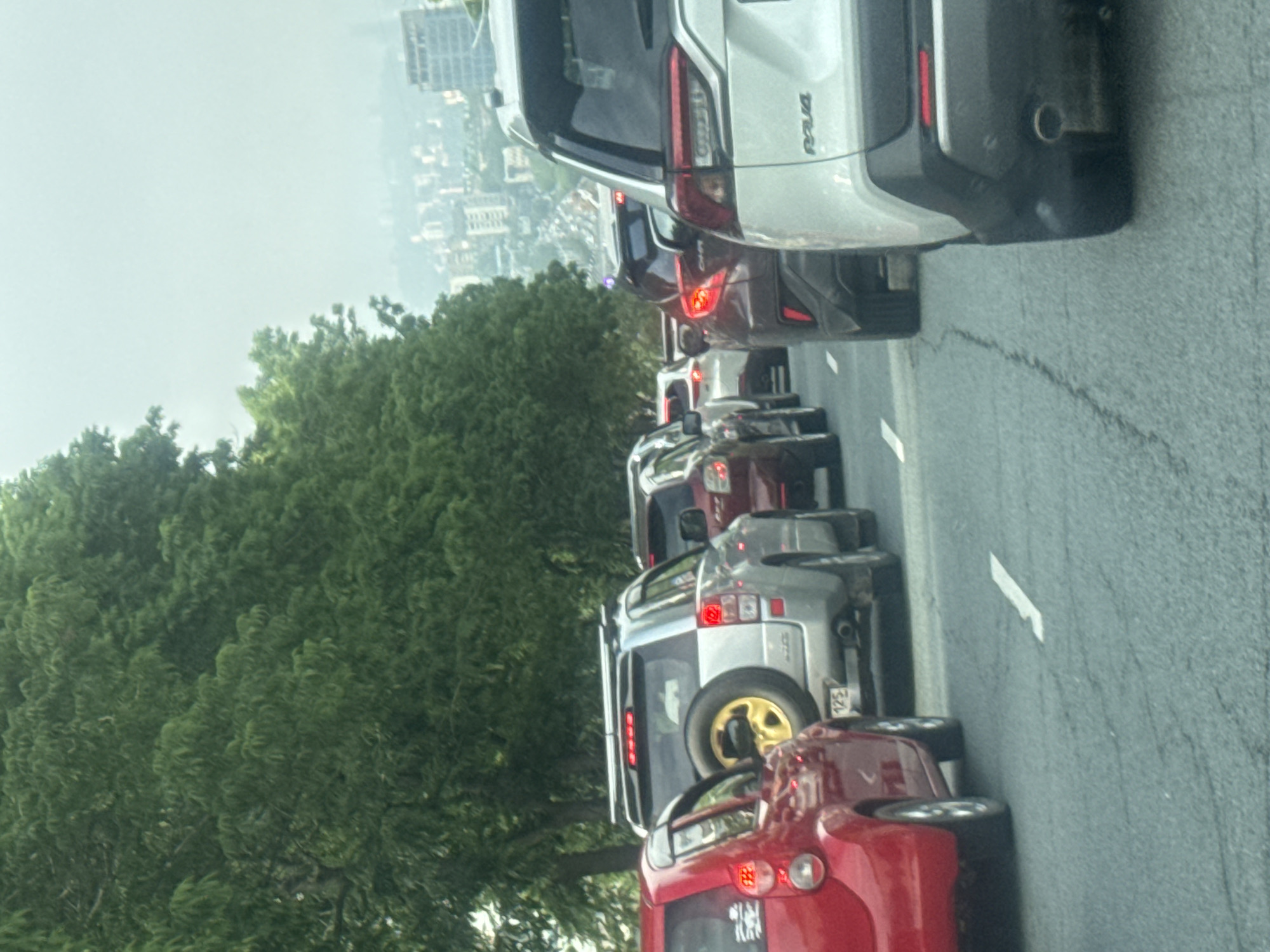 The image shows a congested roadway with multiple cars in a traffic jam. The perspective is from inside a vehicle, looking forward. A red car is visible on the left, followed by a silver SUV with a spare tire mounted on the back. Beyond that, a line of other vehicles, including a dark red SUV and a white SUV, are stopped in traffic. The roadway appears to be paved, with white lane markings visible. Tall green trees line the left side of the road, and a distant cityscape with buildings is visible in the hazy background on the right. The sky is overcast.