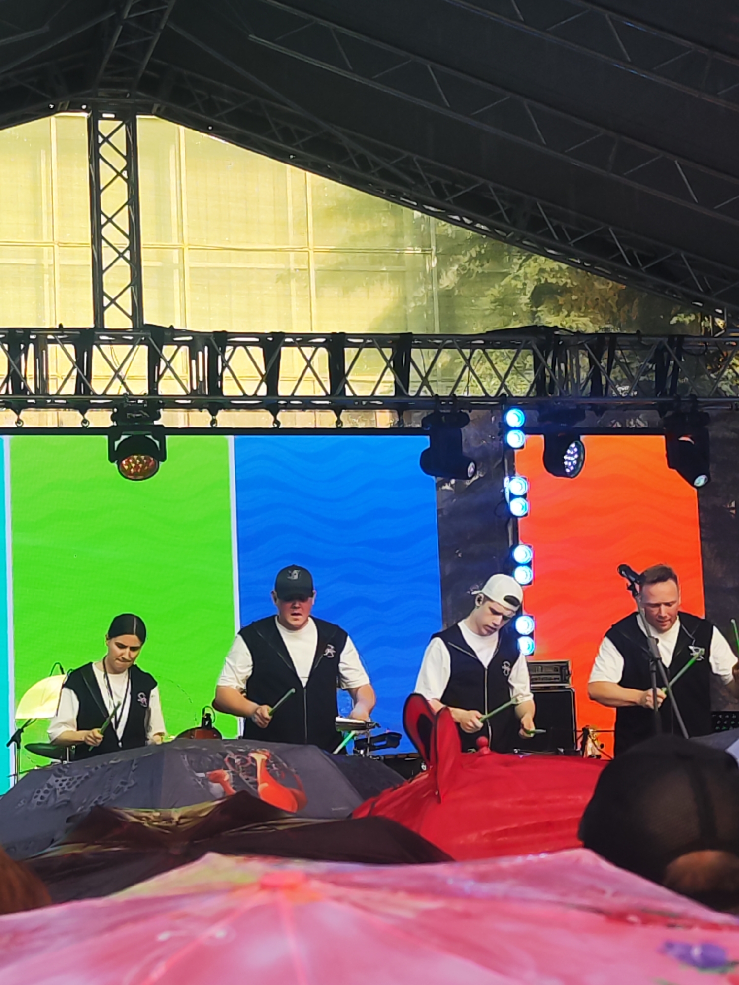 In Chaikovsky, Russia, a musical performance is taking place on an outdoor stage. Four musicians, two men and two women, are positioned behind a row of large, open umbrellas that are decorated with various patterns. The musicians are all wearing dark vests over white shirts. They are holding drumsticks and appear to be playing instruments that are not fully visible, possibly percussion instruments, as suggested by the drumsticks and the electronic pads in front of them.
The stage features a large LED screen backdrop divided into three vertical sections: green on the left, blue in the middle, and orange on the right. Overhead, a metal truss structure supports lighting equipment. In the foreground, the tops of more decorated umbrellas are visible, suggesting the audience is seated or standing behind them. The overall environment appears to be an outdoor event, likely a festival or concert, given the stage setup and the presence of an audience.