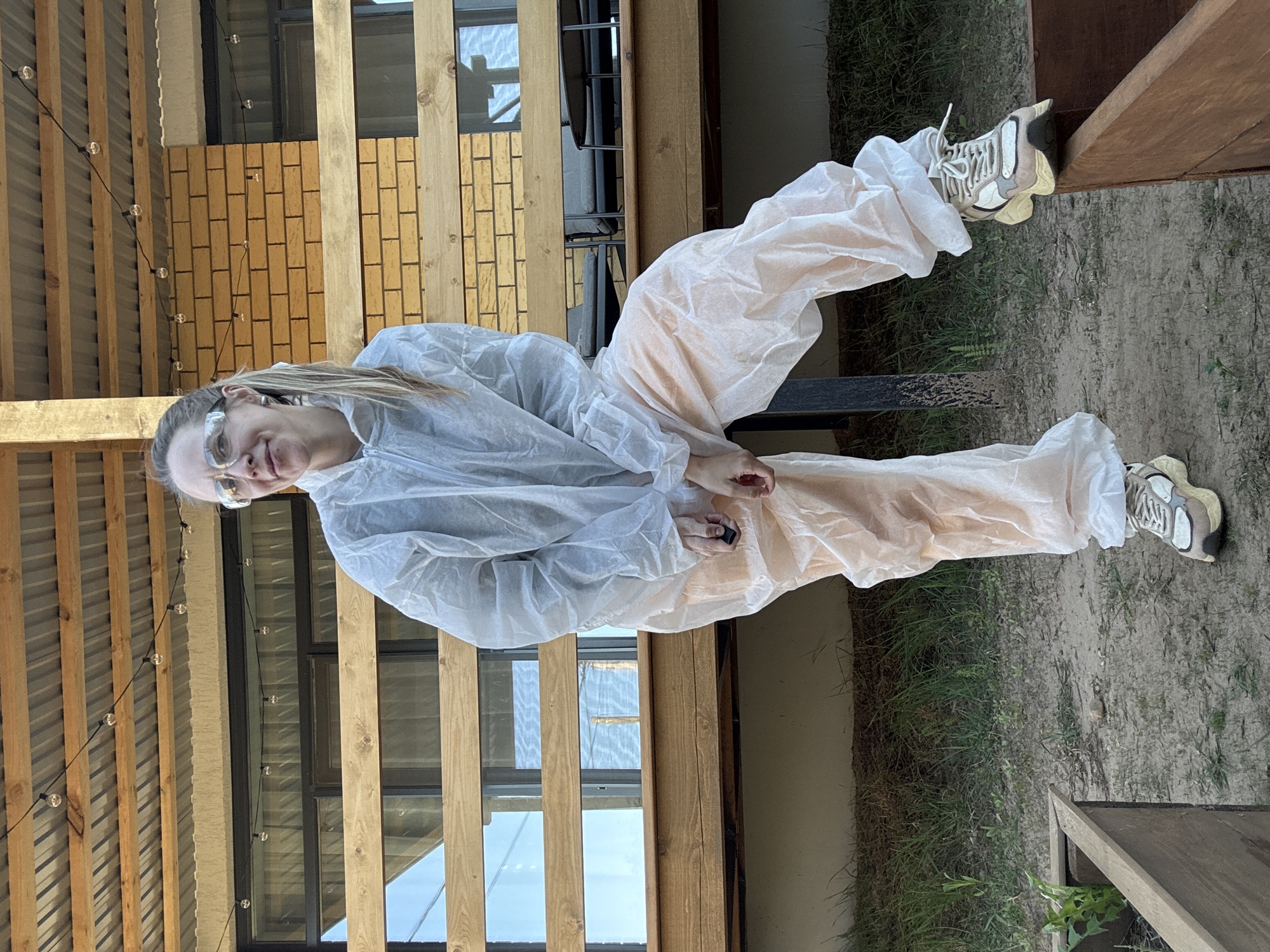A woman wearing a white, disposable protective suit and safety glasses is sitting on a metal structure outdoors. She is holding a small black object in her hand. The woman appears to be smiling. The background shows a building with wooden beams and windows, along with a grass and dirt ground. The image was likely taken in Chaikovsky, Russia.