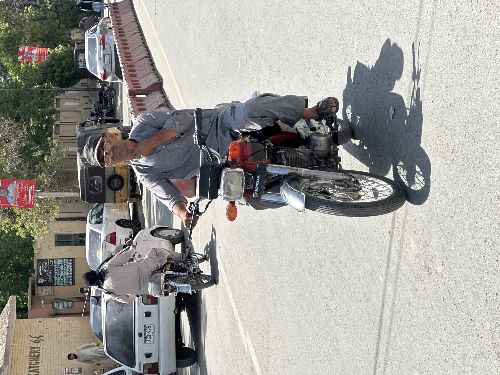 The image depicts a man sitting on a Honda motorcycle on a paved road in Quetta City Tehsil, Pakistan. The man wears a grey shirt, grey pants, sandals, a hat, and glasses. He looks directly at the camera. The motorcycle is red and black. His shadow is cast on the pavement. Behind him are parked vehicles including a white Suzuki van with license plate ACX-925, a white car, and a yellow vehicle. There are buildings and trees in the background. A sign with red writing is visible on a building in the background.