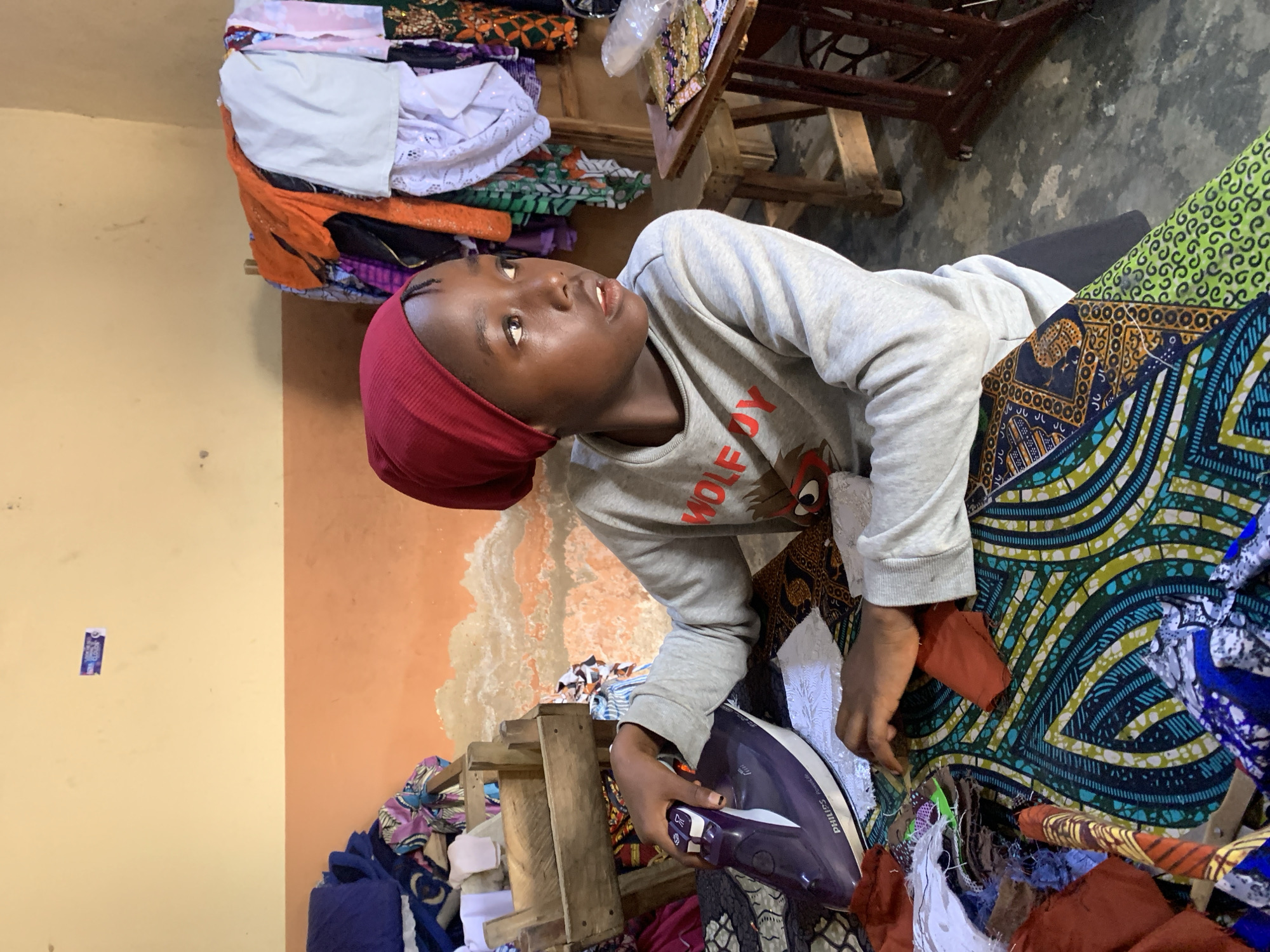 A young person wearing a red headscarf and a grey sweatshirt is ironing a colorful fabric with a purple Philips iron. They are seated at a table cluttered with various pieces of fabric and sewing materials. In the background, clothing hangs on a rack, and a wooden crate is filled with more fabric. The walls are painted in two tones, yellow at the top and orange below, with some visible plaster imperfections. The scene appears to be an indoor space, possibly a workshop or home, in Jos, Nigeria.
