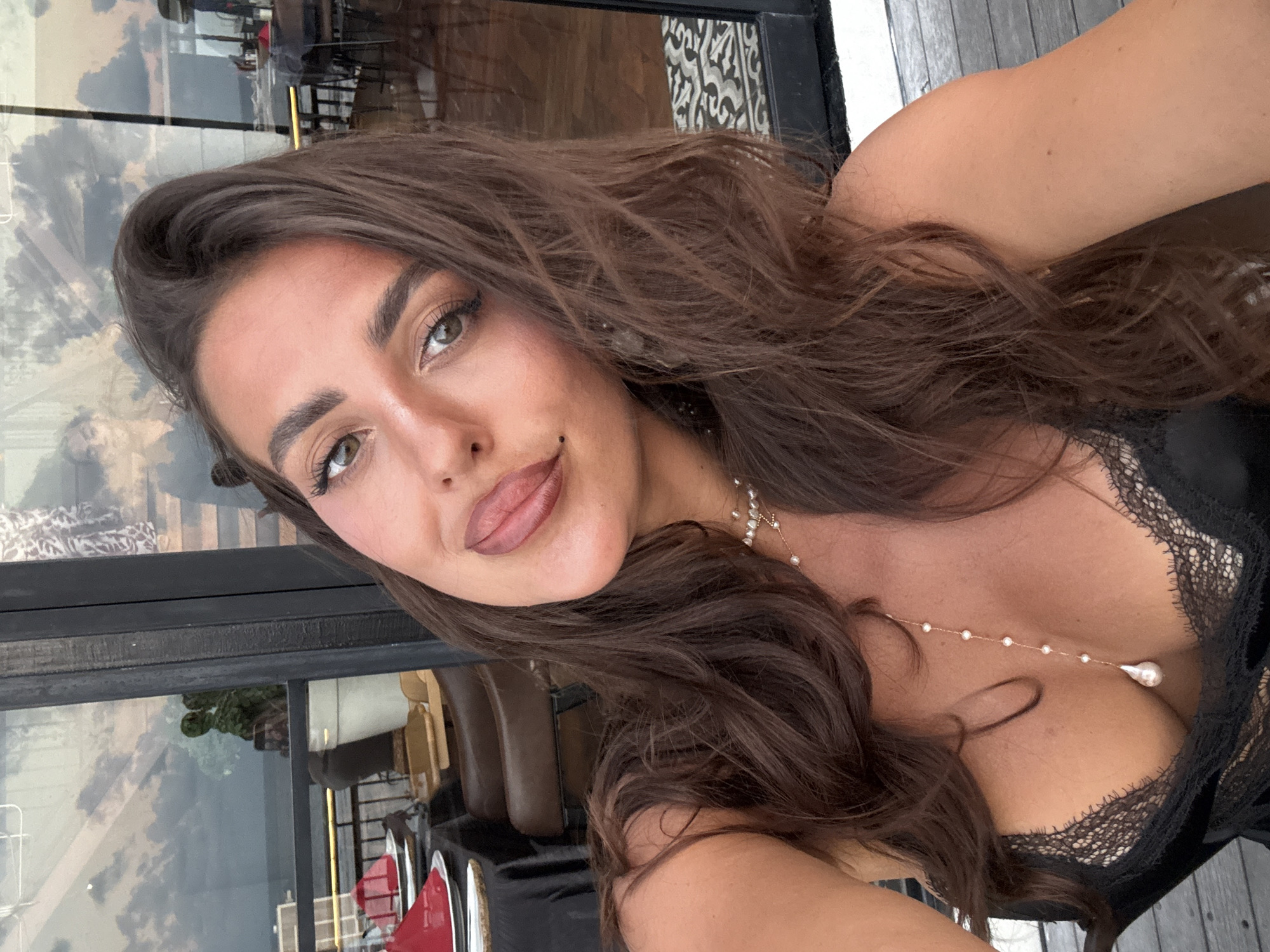 A woman poses for a selfie outdoors in what appears to be a cafe or restaurant setting. The location is identified as Kuta Selatan, Indonesia. She has long, wavy, dark brown hair, light skin, and is wearing a black lace-trimmed top and a pearl necklace. She has light green eyes and a mole on her left cheek. Behind her is a glass wall reflecting the outside environment, including the sky, clouds, and some furniture visible inside and outside the building.