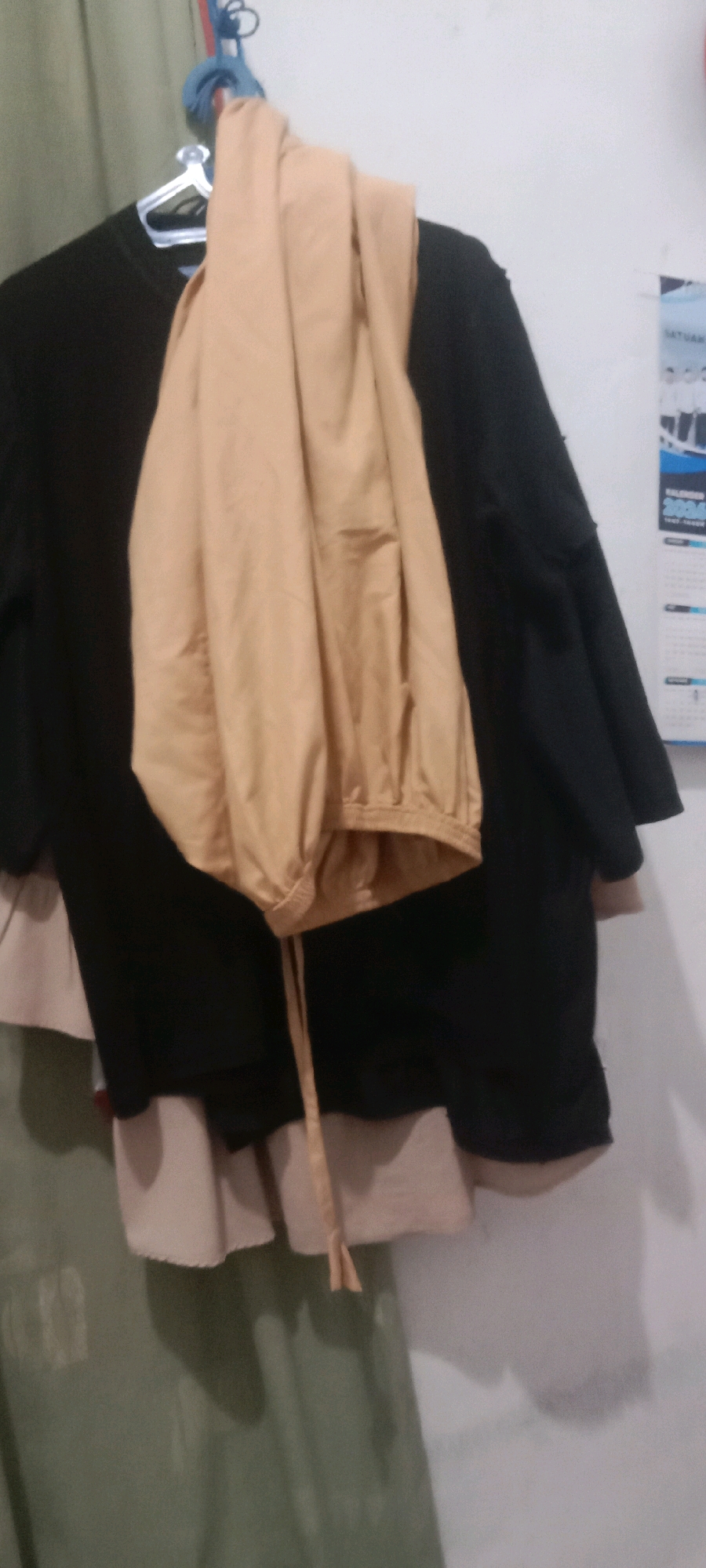 A black shirt and tan pants are hanging on a clothes hanger. The shirt is on the left and the pants are draped over the shirt. The clothes are hanging on a rod, and there is a white wall in the background. There is also a calendar on the wall to the right.