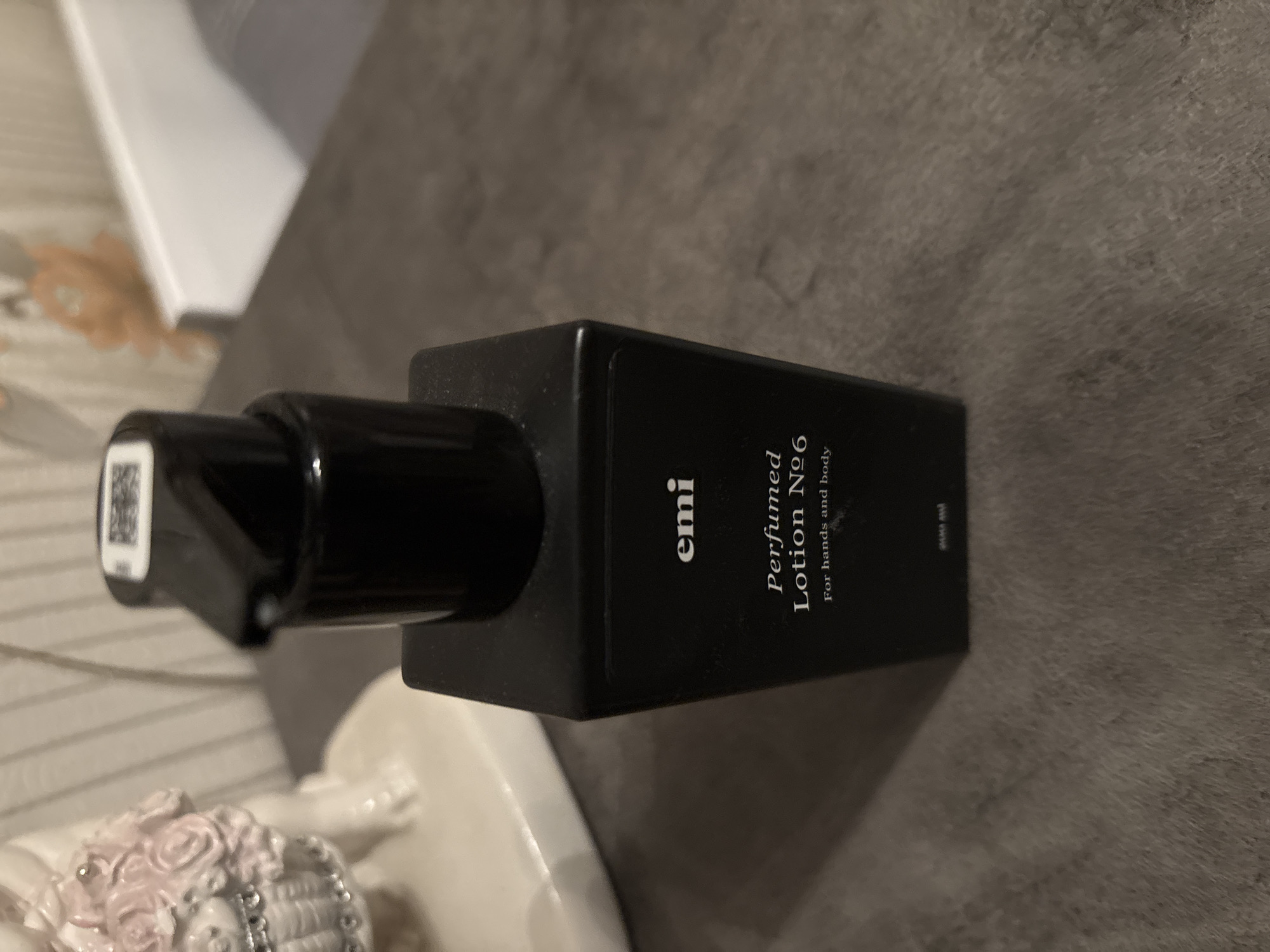 The scene, located in the city of Chernogorsk, Russia as specified, presents an indoor, close-up view of a black, rectangular pump bottle, centrally positioned and slightly angled to the right. The bottle is labeled with "emi" in white sans-serif font at the top. Below this, in white serif font, it reads "Perfumed Lotion Nº6," followed by "for hands and body" in a smaller serif font. At the very bottom, "200 ml" is visible in small white text. The bottle is topped with a black pump dispenser, on which a small white rectangular label with dark, pixilated markings (possibly a QR code or barcode) is affixed. To the left of the bottle, partially visible in the background and out of focus, is a white or cream-colored ceramic figurine. This figurine appears to depict a figure holding a basket adorned with pink roses and small, shiny, possibly silver-colored details. The background behind the bottle and figurine is blurred, revealing vertical striations, indicative of a wall covering such as wallpaper or paneling, along with indistinct lighter-colored forms, including a soft orange hue and a white horizontal line that could be a shelf or furniture edge. The bottle rests on a dark gray, textured surface, possibly a textile or carpet. Lighting suggests an indoor environment, casting subtle shadows.