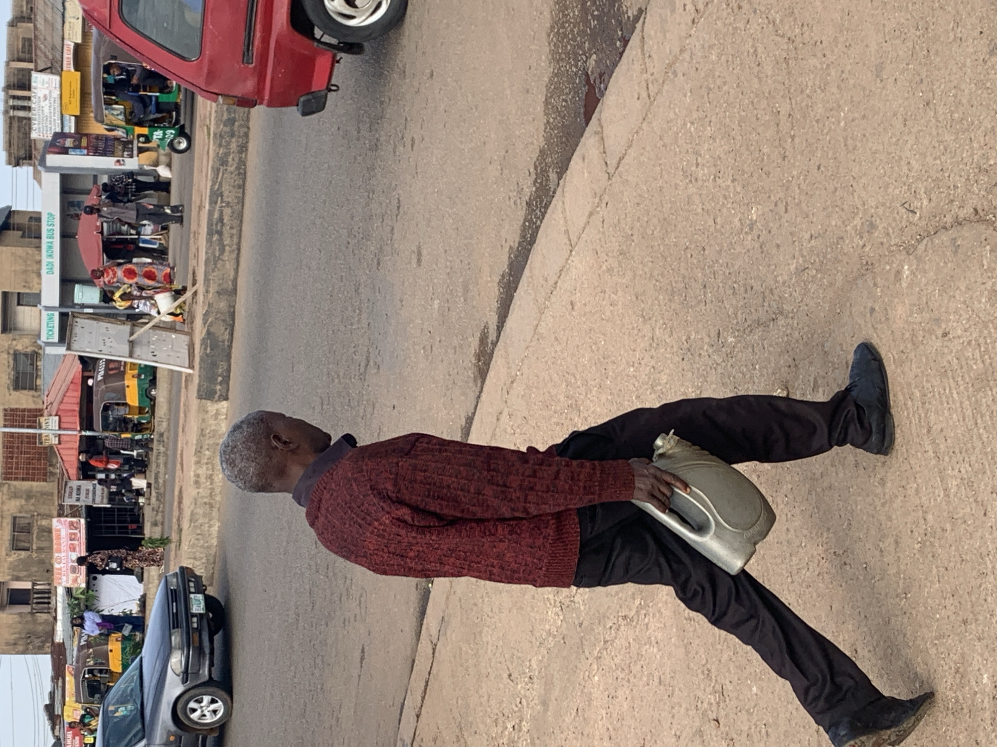 A Black man walks on a sidewalk in Gura Top, Nigeria, carrying a grey plastic jerrycan in his right hand. He wears a maroon, knit sweater over a dark collared shirt and black trousers. His shoes are dark. The sidewalk is cracked and appears dusty. In the background, a grey car is parked on the street, and a red van is partially visible. Beyond the street, there are buildings, including a structure labeled "DADI IKOWA BUS STOP," with people and vehicles, including a yellow and green tuk-tuk, present. The scene appears to be a public area with commercial activity.