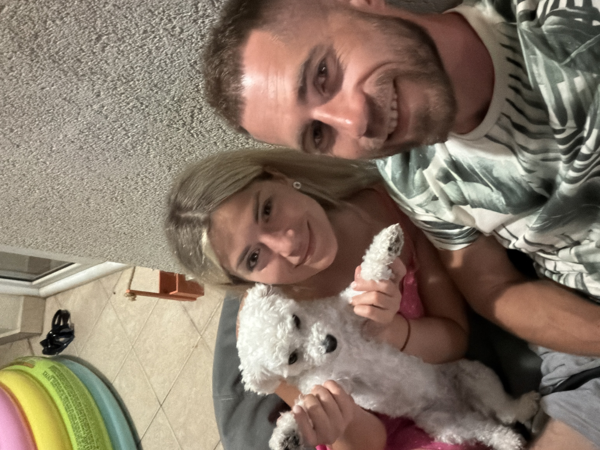 A selfie shows two people, a woman and a man, holding a small white dog outdoors. The woman, with blonde hair and wearing a pink top, is on the left, holding the dog. The man, with short brown hair and a beard, is on the right, smiling. They are seated on a dark cushion with a textured wall behind them. To the left of the frame, a portion of a colorful inflatable ring is visible, resting on a tiled floor. The scene appears to be on a balcony or patio, possibly in Budva, Montenegro, given the architectural style of the wall and the presence of outdoor furniture.