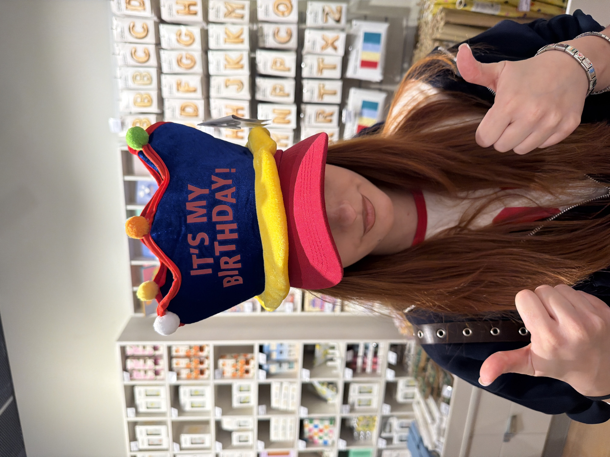 A woman is wearing a red and blue birthday hat with orange text that says "It's My Birthday!" She is giving two thumbs up. She has long red hair and is wearing a black jacket over a white shirt with a red collar. The background shows retail shelves stocked with various items, including letters and cards. The location is a store in Spreitenbach, Switzerland.