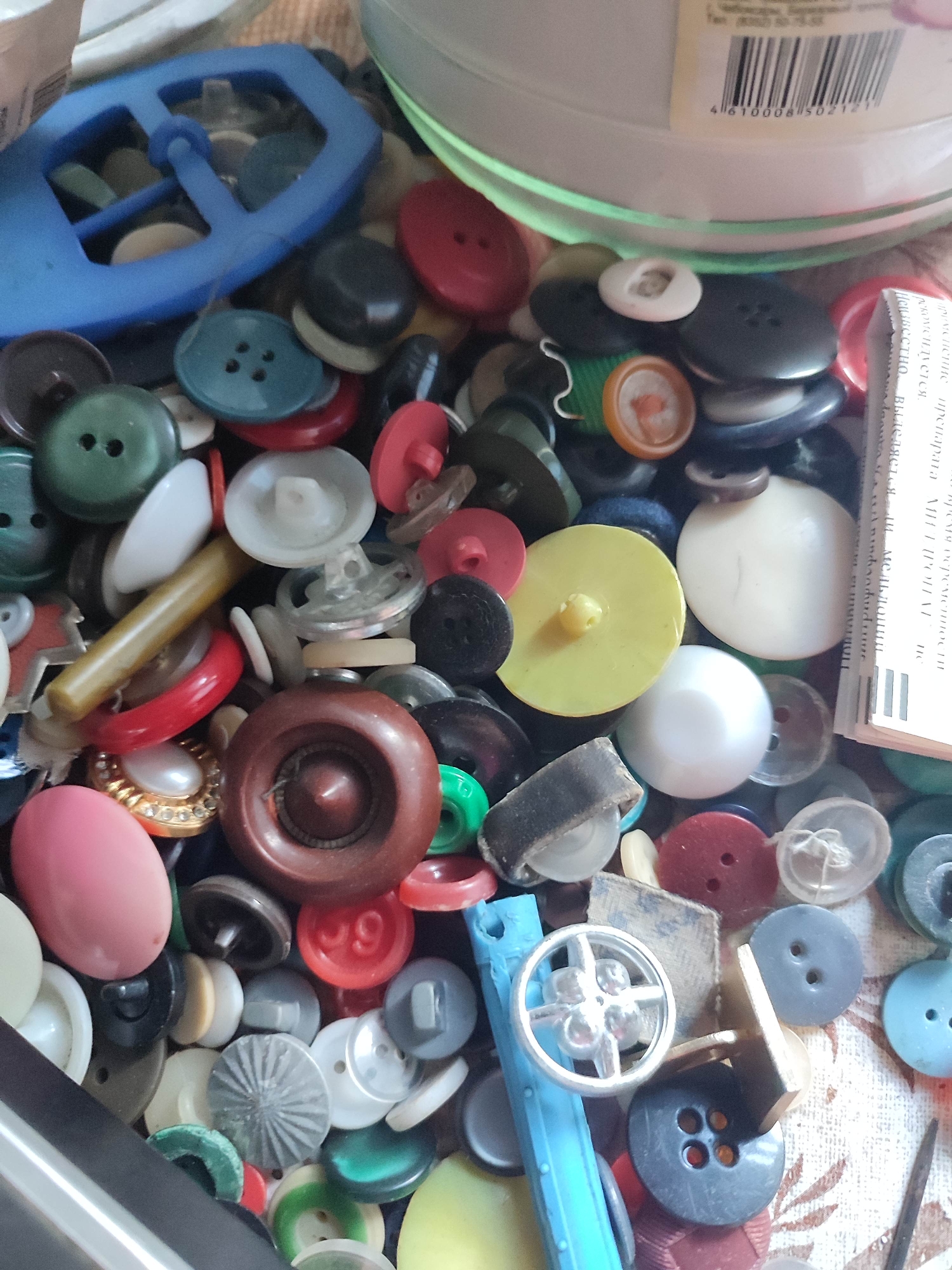 A close-up, overhead shot displays a jumbled collection of buttons and sewing notions. The items are piled on a textured, fabric-like surface, possibly a tablecloth or a sewing mat, with a faint floral pattern visible in the background. The assortment includes various shapes, sizes, and colors of buttons: round, oval, and novelty shapes. There are also a few other sewing accessories, such as a spool of thread, a small metal wheel, and what appears to be a thimble. The overall scene is one of disarray, suggesting a sewing box or craft drawer that has been opened and its contents spilled out. The lighting is natural and somewhat diffuse, casting soft shadows. This image does not contain any information about the city of Gremyachinsk, Russia.