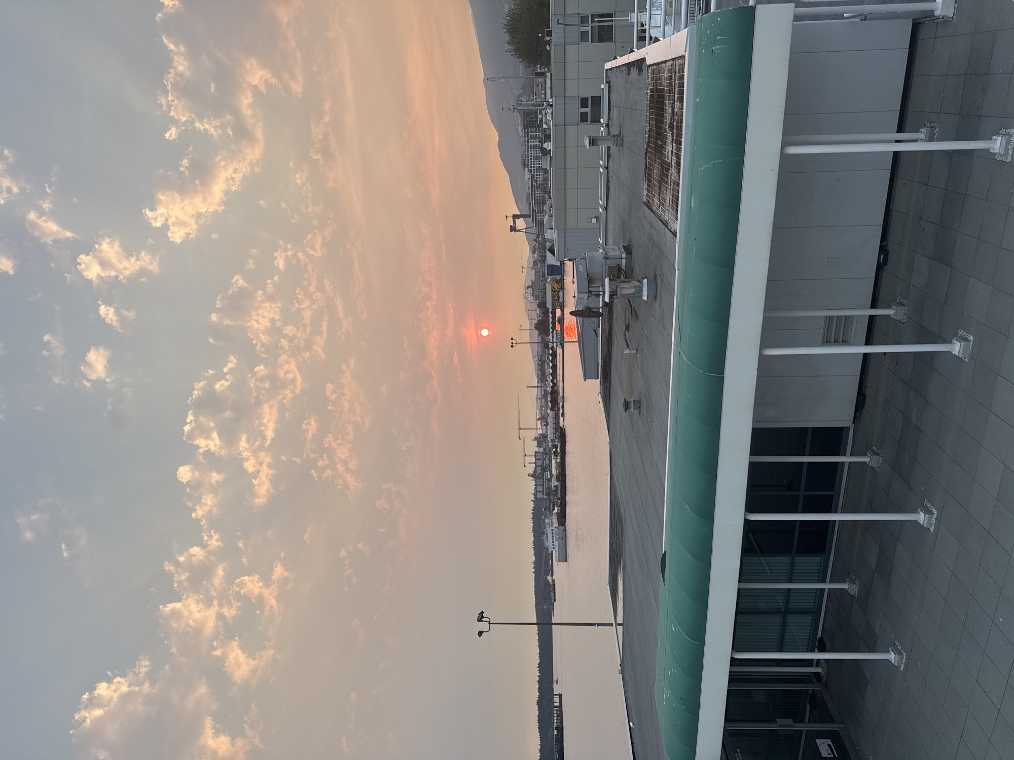 The image captures a hazy sunset over the city of North Vancouver, Canada, likely taken from a vantage point overlooking the waterfront. The sky is filled with diffused clouds tinted in shades of orange, pink, and pale blue, with the sun appearing as a muted, orange-red disc. In the foreground, the flat rooftop of a modern building is visible, featuring a green awning extending over a walkway supported by white columns. Beyond the building, the water of the Burrard Inlet stretches out, reflecting the sunset's colors.
The middle ground is populated by various structures and vessels. A prominent white ferry is docked or moving slowly on the water. Several cranes and industrial buildings line the distant waterfront, suggesting a port or industrial area. Further in the distance, mountains form a hazy silhouette against the sky, typical of the North Shore mountains. The overall atmosphere is one of a subdued, possibly smoke-affected sunset, common during certain seasons in the region due to wildfires. No people are clearly visible in the frame.