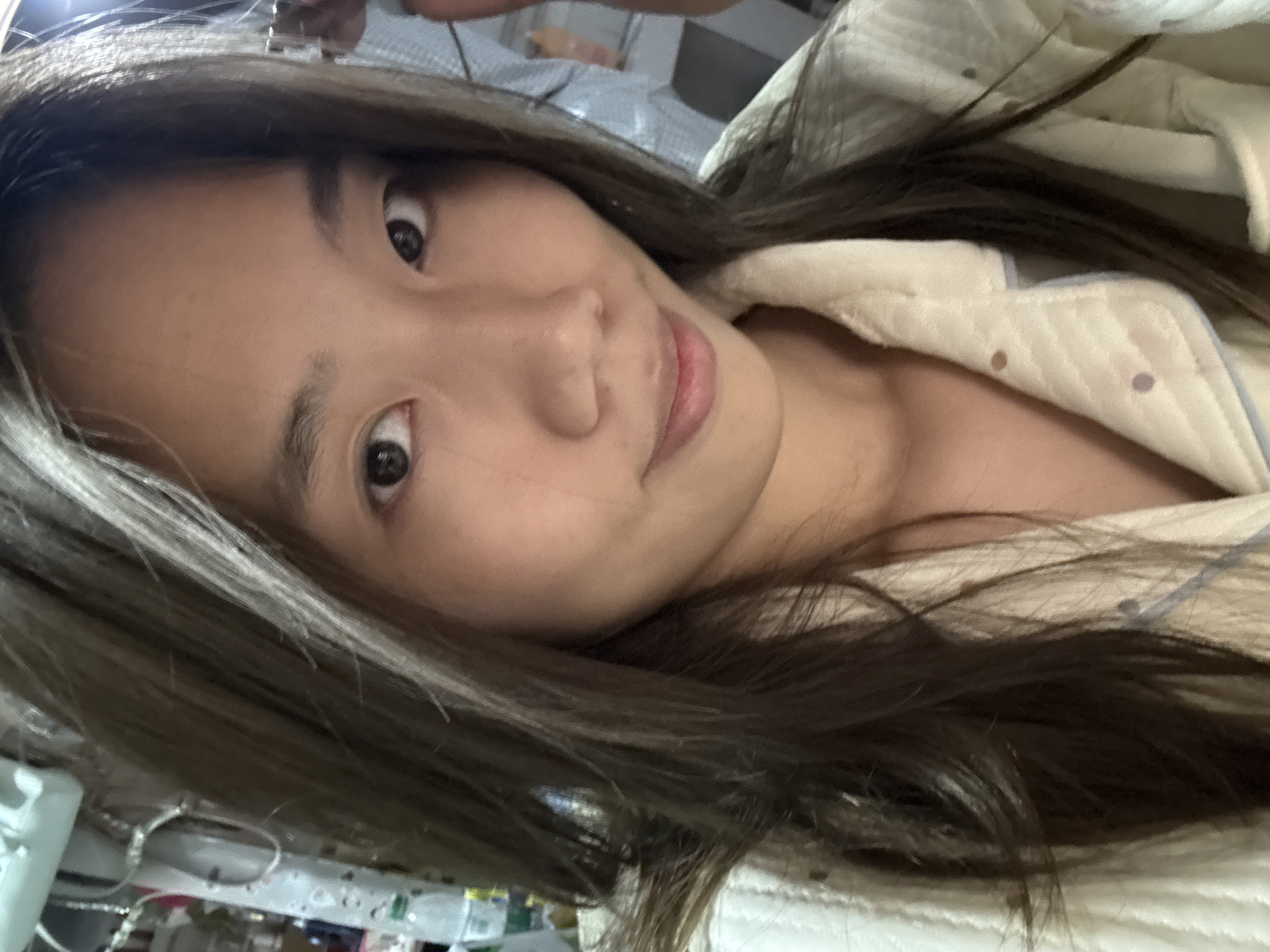 A close-up, rotated selfie shows a young woman with long, dark brown hair that appears to have lighter highlights. Her complexion is light, and her eyes are dark. She is wearing a light-colored, quilted jacket or shirt with small, circular embellishments. The background is blurry and indistinct, but glimpses of what appear to be shelves with items on them and possibly a metal structure can be seen. The location is unknown and cannot be determined from the image. The city of Nan'an District, China, is not discernible from the image.