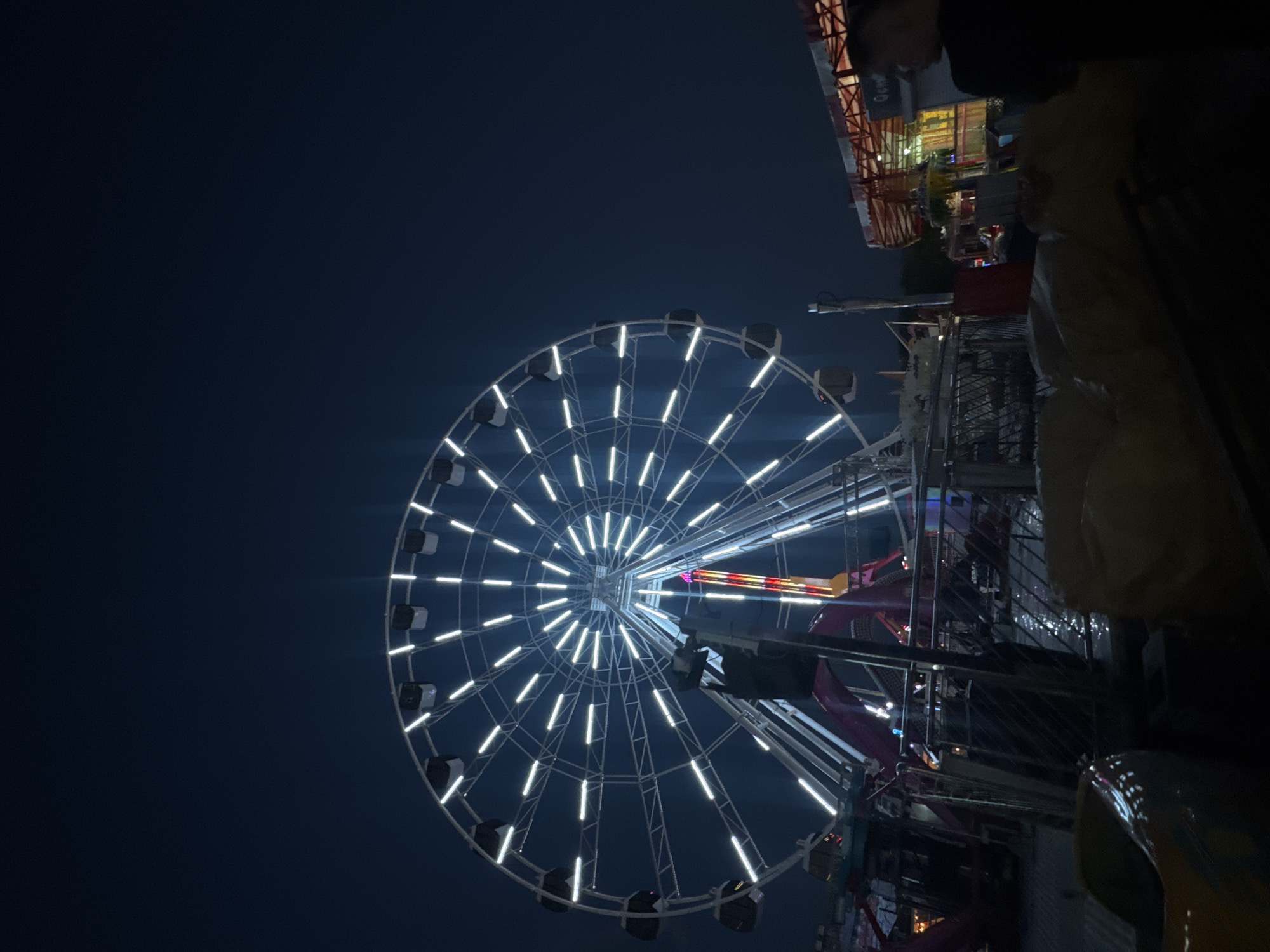 The image shows a Ferris wheel illuminated with white lights against a dark blue sky. The wheel has a center hub with radiating spokes and individual gondolas along the rim. It appears to be nighttime. The photo is taken from a low angle looking up at the wheel. In the foreground, there are parts of another structure, possibly a ride or building, visible with various lights and support beams. The upper right corner shows a partial view of a structure with a red roof. It is likely that this image was taken in a fair or amusement park. While I cannot verify the exact location in Yevpatoriya, Ukraine from the image alone, it is a common feature in amusement parks.