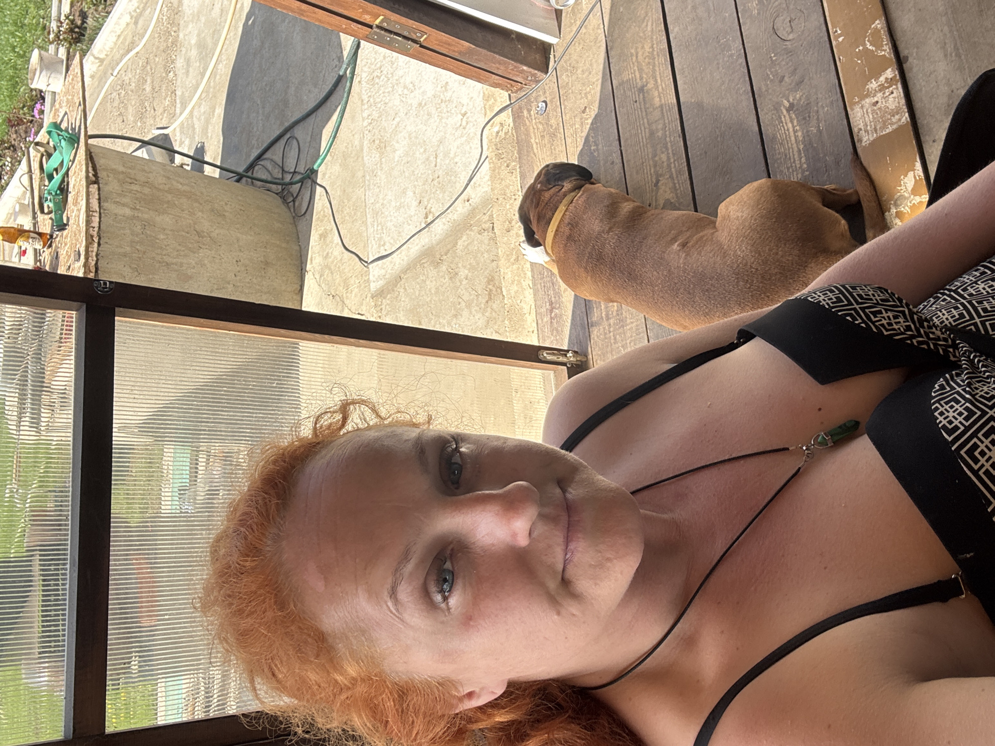 A woman with red, curly hair and light eyes is seen taking a selfie, looking directly at the camera with a neutral to slightly content expression. She is wearing a black top or swimsuit and a necklace featuring a green pendant. The image is oriented sideways, with her face and upper torso occupying the foreground.
In the background, a brown dog, resembling a Boxer breed, with a light-colored collar, is lying relaxed on wooden deck floorboards. Adjacent to the wooden deck is a concrete patio area, where various outdoor items are present. These include green garden hoses, electrical wires, and a large, light-colored, possibly concrete, structure or planter. A translucent, corrugated panel, consistent with a greenhouse or enclosed veranda, frames part of the background, revealing blurred greenery and distant structures. Near one of the green hoses, a small bottle or label is visible with the partial text "Budw," strongly suggesting "Budweiser," along with a distinct red label.
The scene is brightly lit by natural sunlight, indicating a clear day, likely during the morning or afternoon. The overall setting is an outdoor residential area, possibly a backyard patio or veranda, situated in Chaikovsky, Russia, conveying a sense of leisure and domestic tranquility.