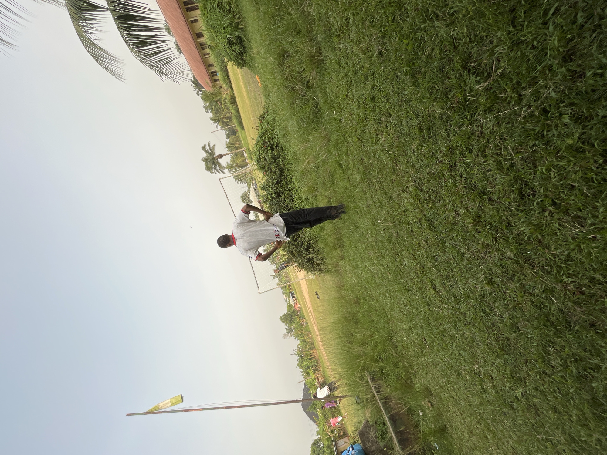 The image depicts a grassy field in Ikot Ekpene, Nigeria, under an overcast sky. A person wearing a white shirt and dark pants stands in the midground. A goalpost is visible in the background, suggesting a sports field. Palm trees and buildings are also present in the background. The grass appears overgrown.