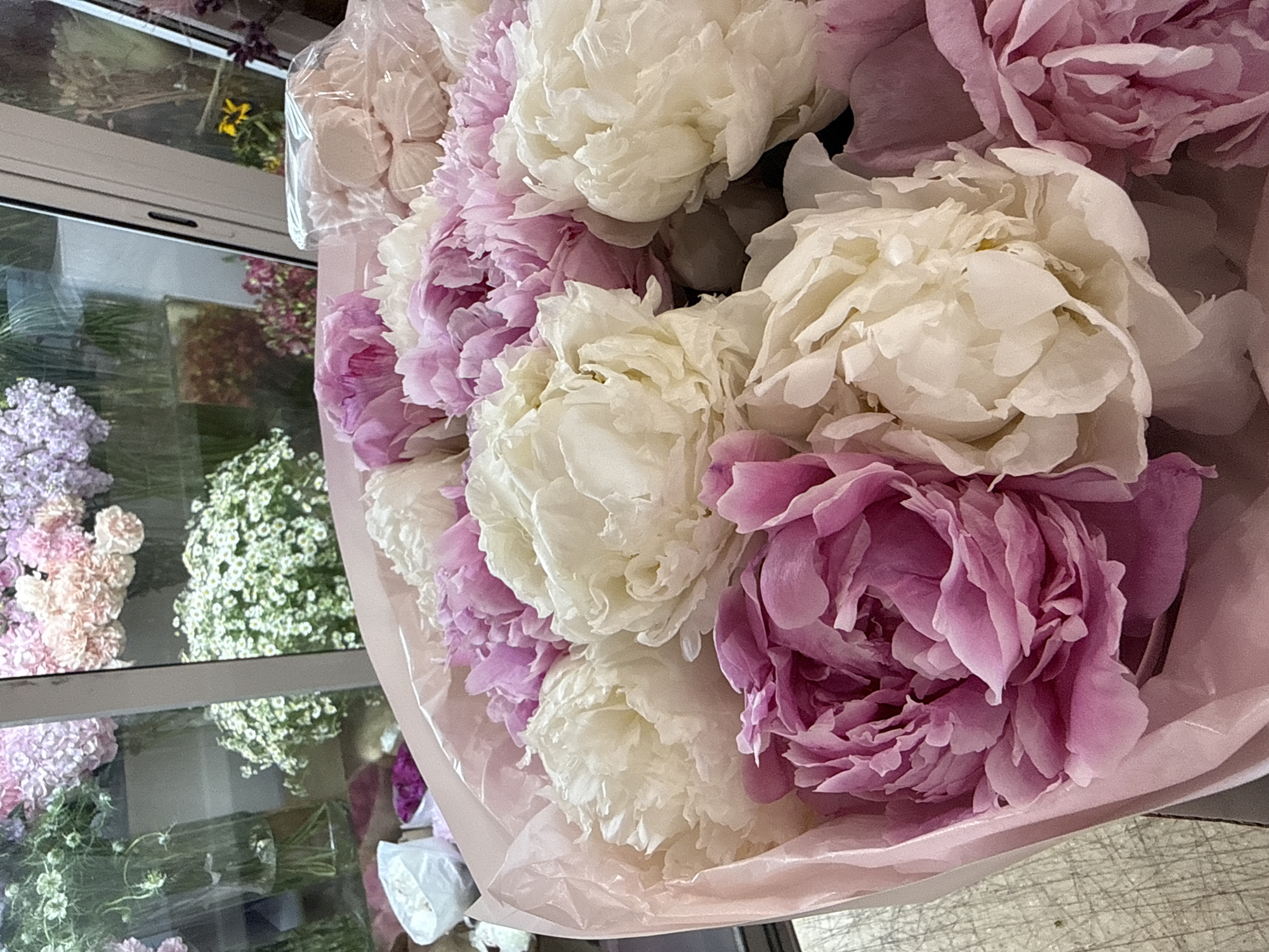 A close-up, overhead shot shows a bouquet of pink and white peonies. The flowers are large and full, with many ruffled petals. They are wrapped in pink paper. In the background, the bouquet is reflected in a window, showing more flowers, including lilac, white baby's breath, and pink carnations. There are also some pink marshmallow-like candies in a clear plastic bag visible on the left side of the frame. The lighting suggests it is daytime. The overall mood is delicate and romantic. There is no visible text.