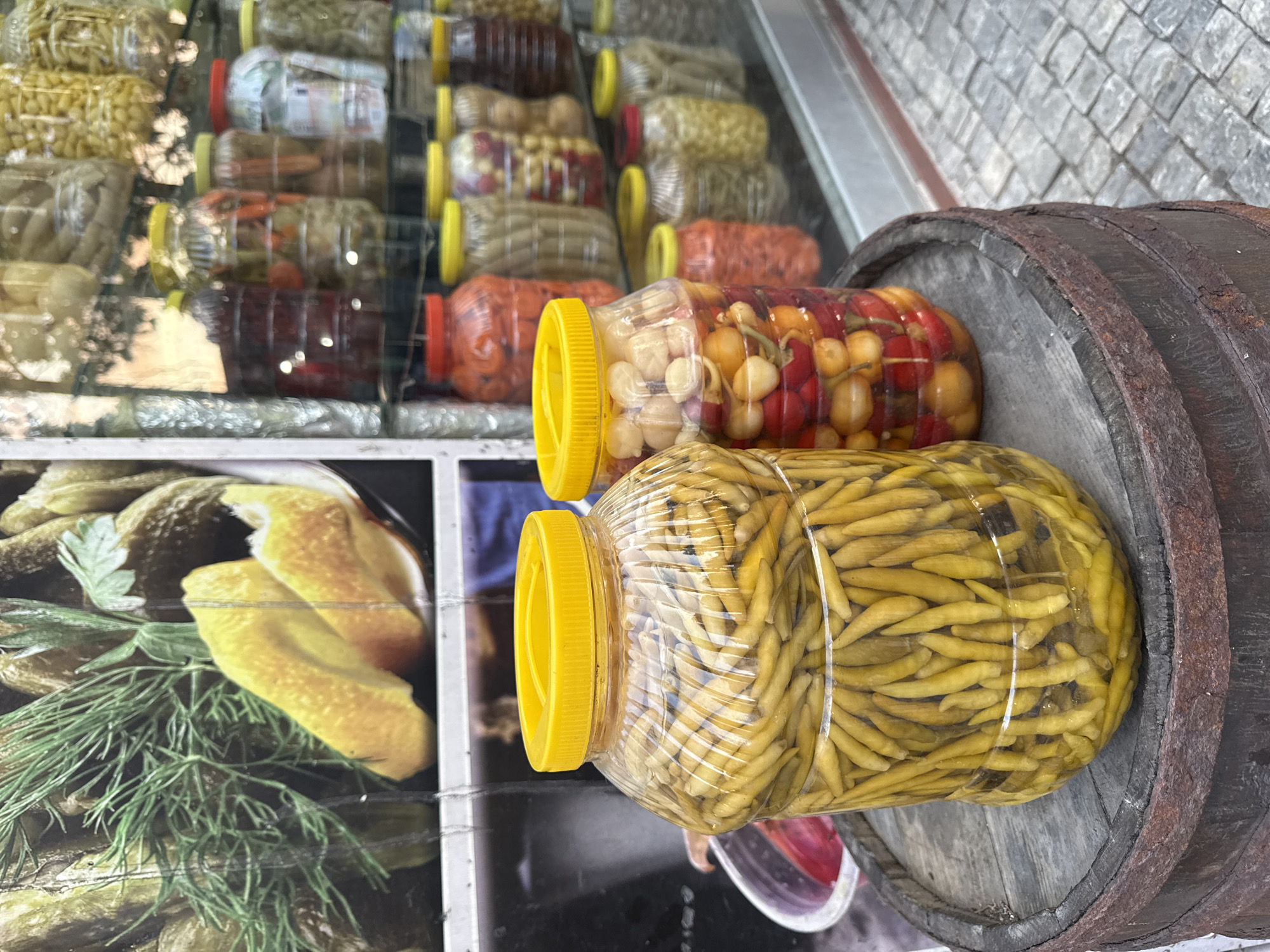 In Kadıköy, Turkey, a weathered wooden barrel sits on a cobblestone street, serving as a display for two large plastic jars of pickles. The jar on the left is filled with long, thin, pale yellow peppers, while the jar on the right contains a colorful mix of small red and yellow round vegetables, possibly cherry tomatoes and small peppers, alongside some chili peppers. Behind the barrel, a storefront window showcases a variety of pickled goods in glass jars, ranging in color from deep red to pale yellow and brown. The storefront display is illuminated, and reflections of the street and surrounding environment are visible on the glass. The overall scene suggests a traditional market or store selling preserved foods.