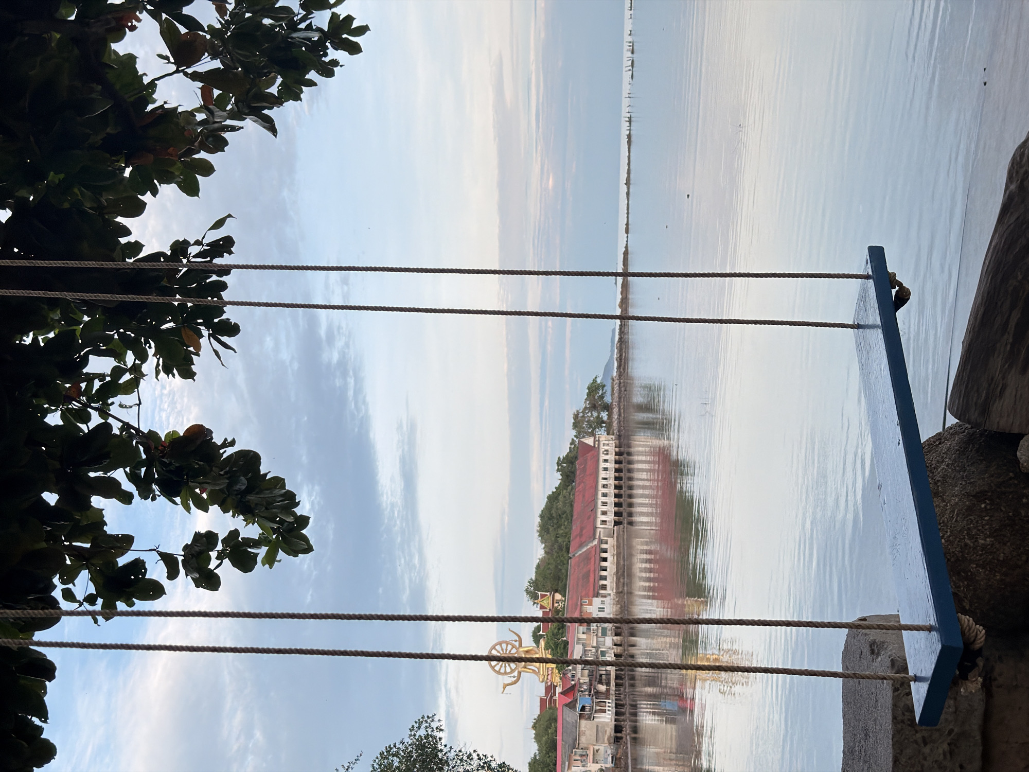 In the foreground, a blue wooden swing hangs from thick ropes attached to a tree branch overhead, suspended over large stones and logs. The swing is empty. In the midground, a calm body of water reflects the sky and a row of buildings with red tiled roofs. To the left of the buildings, a large golden statue of a figure with a wheel of dharma atop its head is visible. The background consists of a serene sky with scattered clouds. The scene appears to be located in a coastal area of Ko Samui District, Thailand, likely in proximity to a temple or cultural landmark given the statue. The time of day seems to be either early morning or late afternoon, judging by the soft light and tranquil atmosphere. There are no people visible in the image.