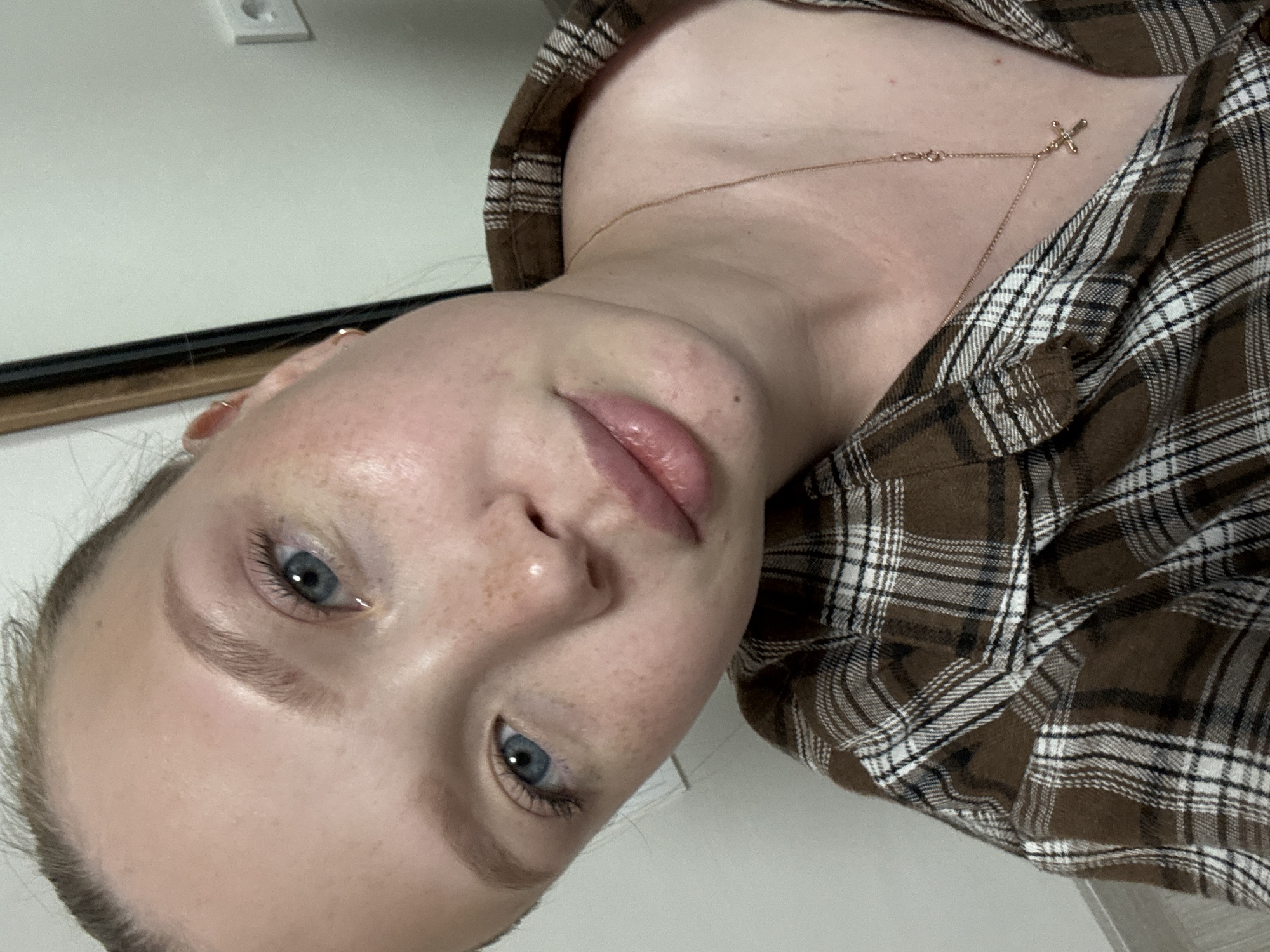 A close-up selfie shows a young woman with blonde hair pulled back from her face, revealing light blue eyes and freckles across her nose and cheeks. She is wearing a brown and white plaid collared shirt and a delicate rose gold necklace with a small cross pendant. Her lips are slightly parted, and her gaze is directed at the camera. The background is a plain white wall, with a corner of a dark-framed mirror and a white electrical outlet visible on the right side. The lighting is soft and natural. The image appears to have been taken indoors, likely in a residential setting in Chaikovsky, Russia.