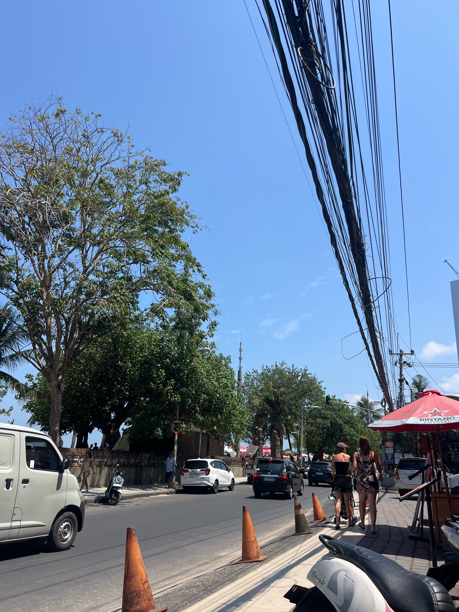 The image captures a street scene in Kuta, Indonesia, on a bright, sunny day. A white van is parked on the left side of the road, facing left. The road itself is paved, with several vehicles, including a white SUV and two black cars, driving or parked. Traffic cones are placed along the side of the road, indicating possible construction or lane closures.
A sidewalk runs parallel to the road, with a few people walking. Two women are walking on the sidewalk, one pushing a stroller. A parked scooter is visible near the sidewalk. A red umbrella with "BINTANG CRYSTAL" branding is open, suggesting a vendor or establishment nearby.
The background features lush green trees and palm trees, with a clear blue sky overhead. A complex network of electrical wires hangs above the street. A low brick wall runs along the side of the road, separating it from the area with trees and some figures in the distance. The overall impression is a bustling street scene in a tropical urban environment.
