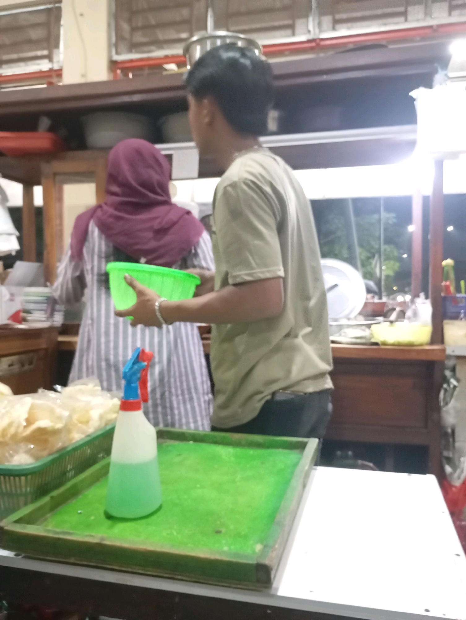 In Babelan, Indonesia, two individuals are present at a food stall. A person wearing a purple hijab and a striped shirt is holding a green plastic bowl. To their right, a person in a light-colored t-shirt and dark pants stands with their back to the camera, facing away from the food stall. In the foreground, a green tray contains a spray bottle with a blue and red nozzle and green liquid, and a basket filled with light-colored snacks. The stall appears to be set up outdoors or in an open-air market.