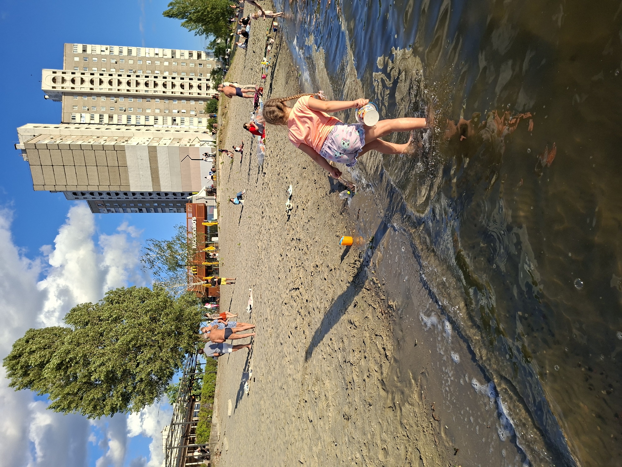The image captures a sunny day at a sandy beach, likely in a city with a waterfront. In the foreground, a young girl with braided hair stands in shallow water, holding a bucket in her right hand and a stick in her left. She is wearing an orange t-shirt and patterned shorts. Gentle waves lap at the shore, creating a foamy edge. Further back on the sand, several people are scattered, some walking, some sitting, and some playing. A group of three adults are standing near a large tree. In the background, a cluster of tall, multi-story buildings, characteristic of urban architecture, dominate the skyline. A McDonald's restaurant with its distinctive yellow arches is visible, suggesting a commercial area adjacent to the beach. The sky is blue with scattered white clouds, and the sunlight casts long shadows across the sand. The overall scene depicts a leisurely day at a public beach.