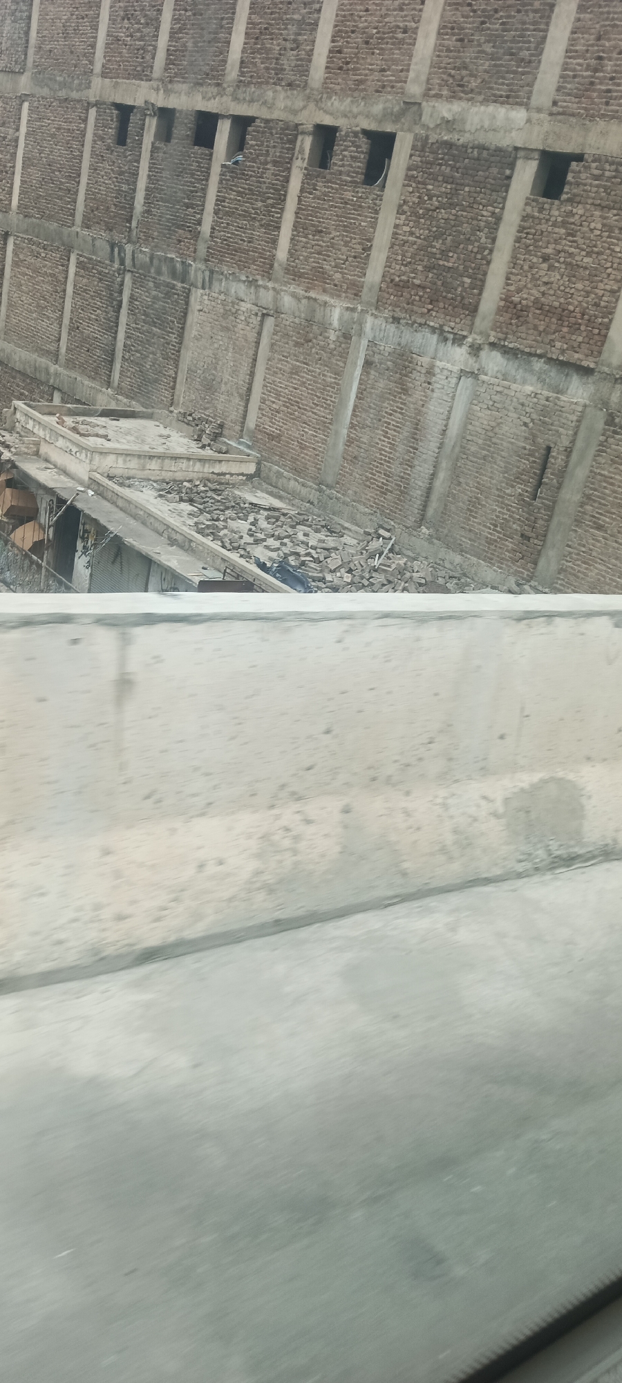 The image shows an unfinished or damaged multi-story brick building in Alam Khan Kalay, Pakistan. The building appears to be constructed with exposed brick walls and concrete pillars. One section of the building, visible in the foreground of the main image and more clearly in cropped images, shows a partially collapsed roof or upper level littered with debris, including bricks and what appears to be blue plastic sheeting. There are numerous openings or windows visible on the different levels of the building, some of which are dark and appear empty, while others may contain objects or remnants. The lower part of the image is dominated by a blurred concrete barrier and road, suggesting the photo was taken from a moving vehicle or elevated position overlooking the scene. There are no people visible in the image. The overall impression is one of disrepair or ongoing construction with significant material accumulation on the ground.