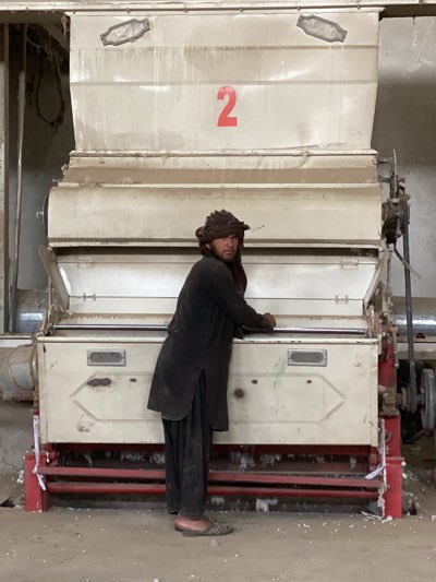 A man stands in front of a large, industrial machine inside a building, likely a factory. The machine is predominantly off-white and appears to be old and used, with a red number "2" painted on its upper section. The man is dressed in dark-colored, traditional clothing. He is leaning slightly on the machine, with his hands resting on its edge. He has a dark complexion and his head is wrapped in a cloth. The factory setting suggests an industrial area, possibly in Kandahar, Afghanistan. The floor is concrete, and the overall lighting is dim, adding to the sense of a utilitarian, working environment.