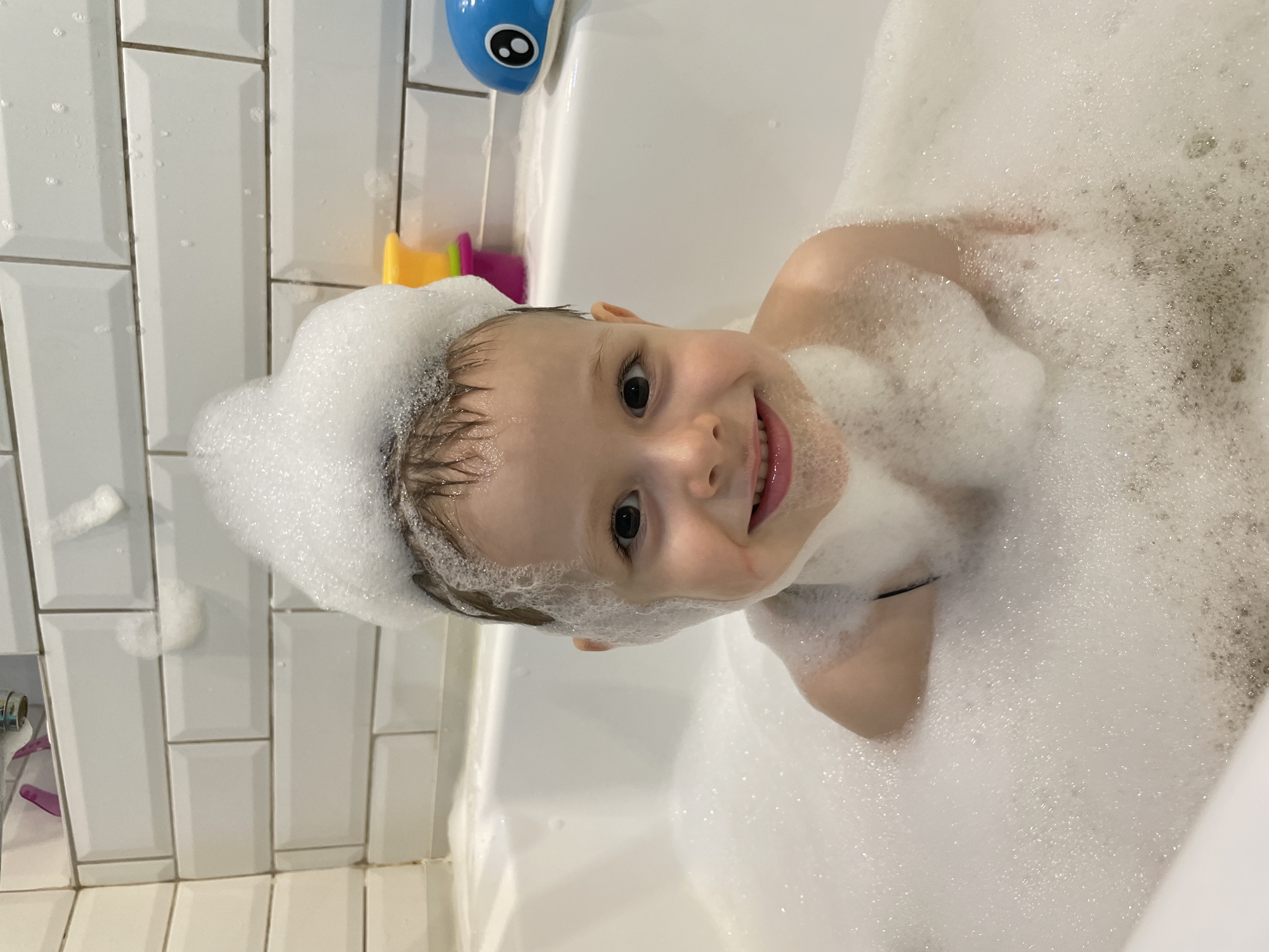 A young child with a head full of bubbles smiles at the camera while sitting in a bathtub filled with foam. The child is wearing a black strap. To the right of the child are yellow and pink bath toys and a blue whale-shaped toy. The bathtub is white with white tiled walls behind it. The image appears to be taken in a residential bathroom.