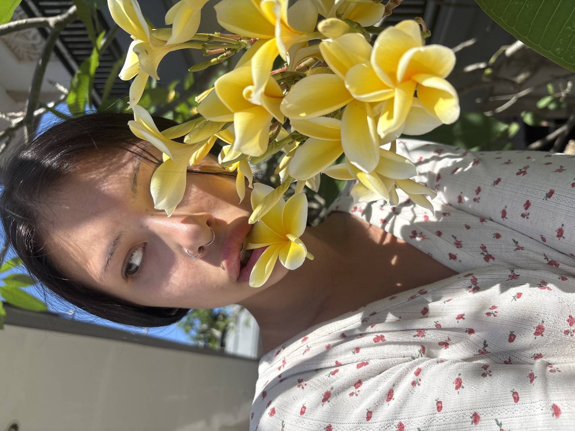 A medium shot shows a young woman in the foreground, holding a yellow frangipani flower in her mouth. She is wearing a white, short-sleeved, V-neck top with a small red floral pattern. Her dark hair is pulled back, and she has a nose ring. The background is blurred, showing hints of a building and foliage under a bright sky. The scene appears to be outdoors, possibly in a garden or courtyard.