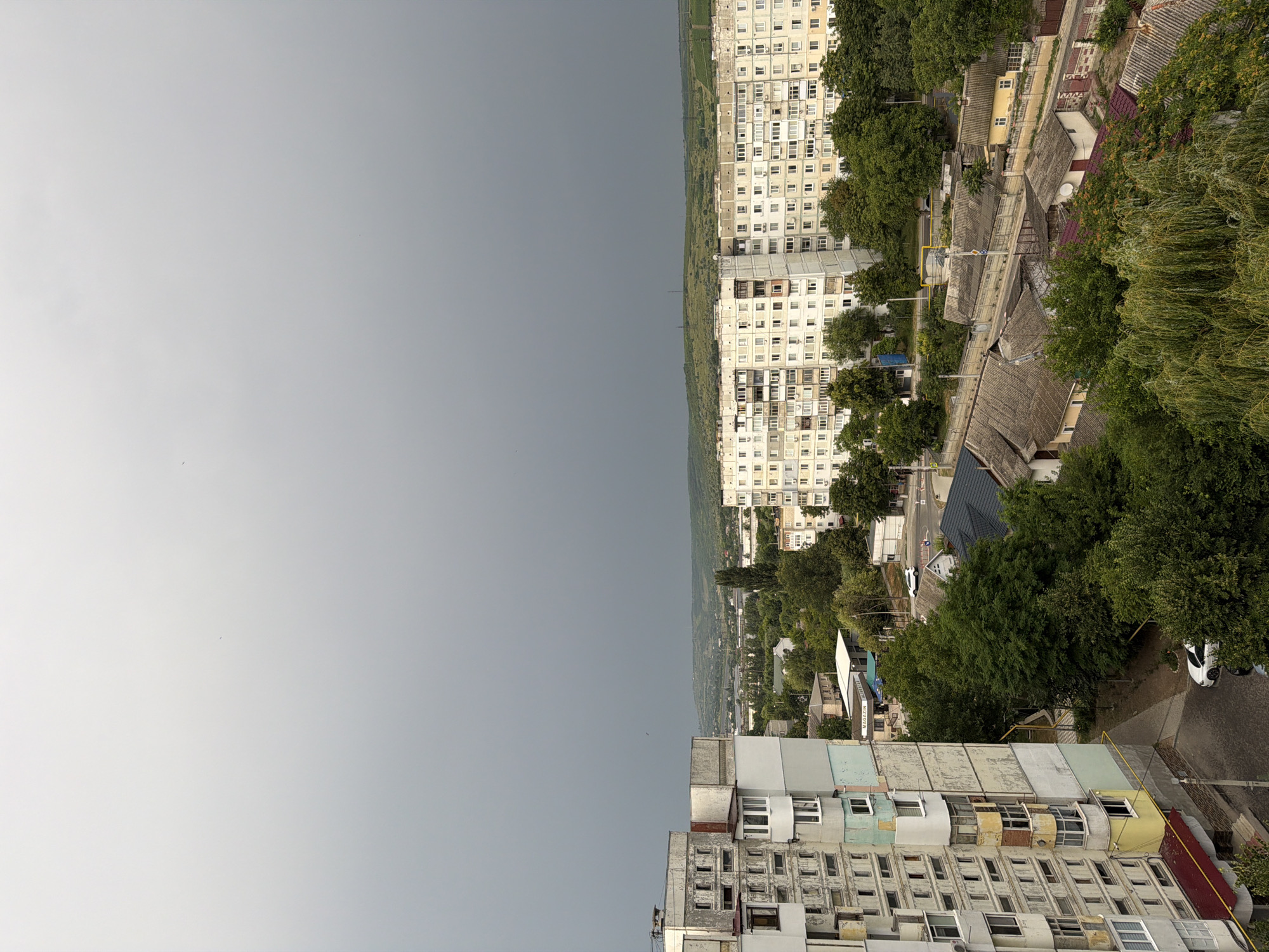 This is an aerial photograph of a city scene in Strășeni, Moldova, under a heavily overcast sky, suggesting imminent or ongoing rain. The foreground features the side of a weathered, multi-story residential building with a distinct concrete facade and numerous balconies. To the right and in the midground, a long, pale apartment block stretches horizontally across the frame, its windows reflecting the dull sky. Between the residential buildings and further into the distance, a mixture of lush green trees and smaller, older houses with grey, tiled roofs are visible. A road with a few parked cars, including a white car, winds through the residential area. The background consists of rolling hills covered in greenery. The overall impression is of a lived-in urban environment during a grey, possibly humid, day.