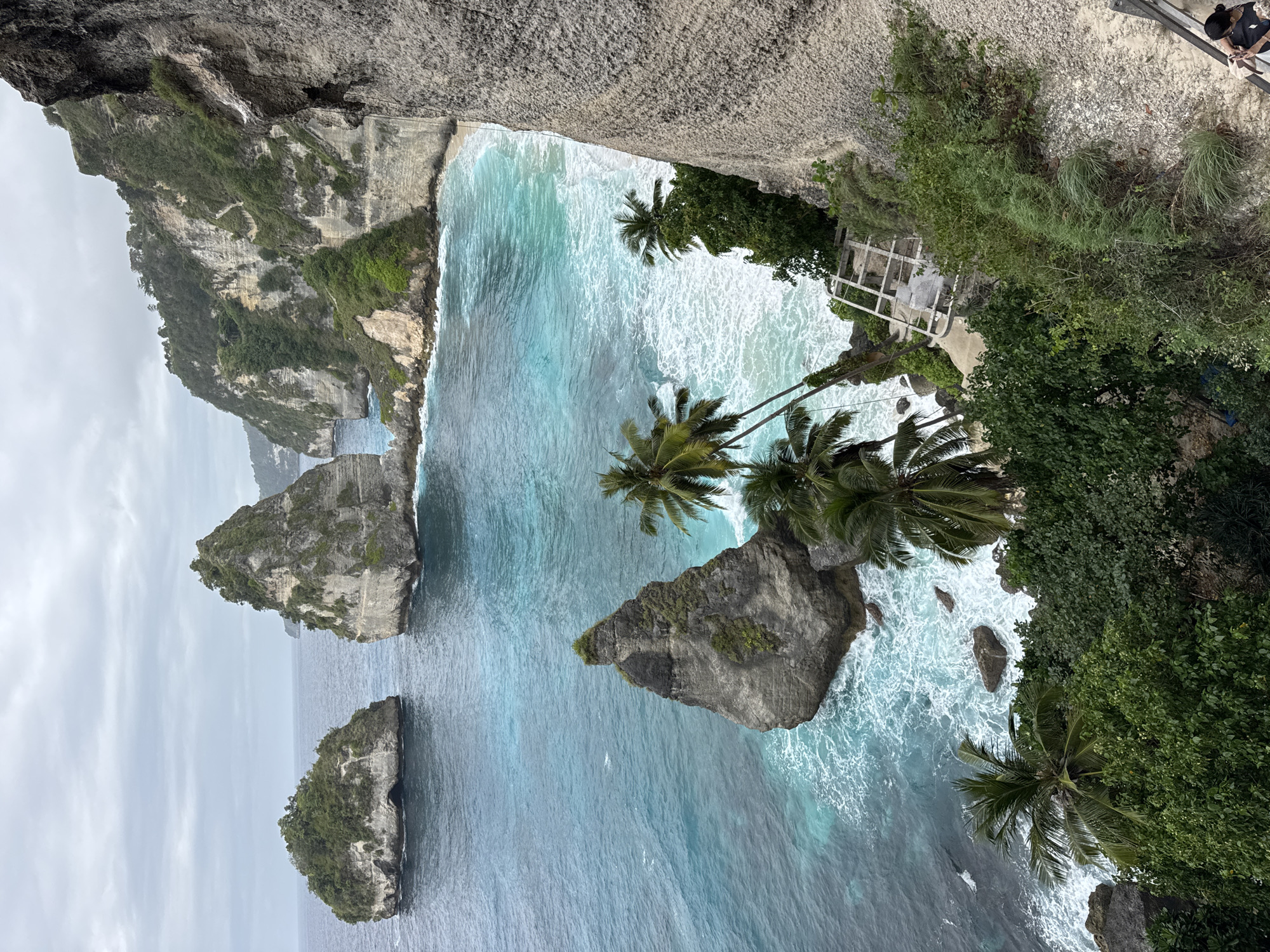 This image depicts a scenic coastal view of Nusa Penida, Indonesia, likely at Diamond Beach or a similar viewpoint overlooking the ocean and rock formations. The scene is dominated by the turquoise waters of the ocean meeting a sandy beach, framed by towering cliffs covered in lush greenery. Several large, rocky outcrops rise from the sea, some topped with vegetation. Palm trees are scattered along the cliff edge and on the beach. The sky is overcast, suggesting a cloudy day. A white structure, possibly a viewing platform or remnants of a building, is visible on the cliff face. In the lower right corner, a person wearing a dark top and light-colored bottoms is seated on what appears to be steps or a pathway leading down from the viewpoint. Another person is partially visible further down the steps. The water exhibits a lively surf with white foam washing onto the shore.