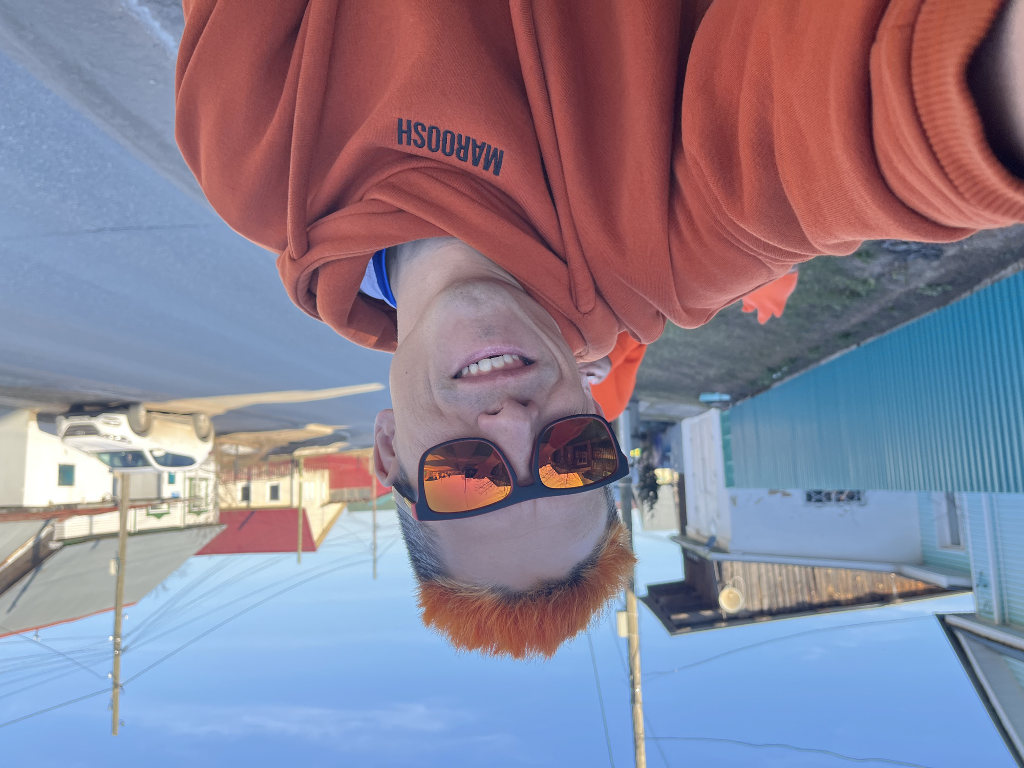 The image is a selfie of an adult male, taken with the camera inverted, making him appear upside down against a clear sky. He is smiling broadly, revealing his teeth. He wears a rust-orange hooded sweatshirt with the brand name "MAROOSH" printed in black on the chest, visible upside down in the current orientation. His hair has distinctive bright orange-red tips, contrasting with darker roots. He is wearing black-framed sunglasses with orange reflective lenses that mirror the surrounding buildings and sky.
The background suggests an outdoor urban or suburban setting, likely a street in Chaikovsky, Russia. Visible elements include several residential-style buildings with varied architecture and roofing (some light-colored, one with a red roof, and another with light blue corrugated metal siding). Utility poles and numerous overhead wires crisscross the scene. A white car is partially visible further down a paved road. The bright light and clear blue sky indicate a sunny day, possibly morning or late afternoon. The man's cheerful expression contributes to a positive overall mood.