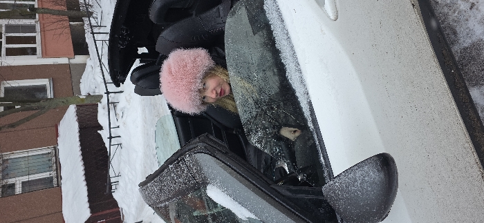 A woman with blond hair and a pink fur hat sits inside a white car. Snow coats the car's exterior, including the open door and windows. Outside the car, a building with snow-covered surfaces is partially visible. The setting suggests a cold, snowy environment.