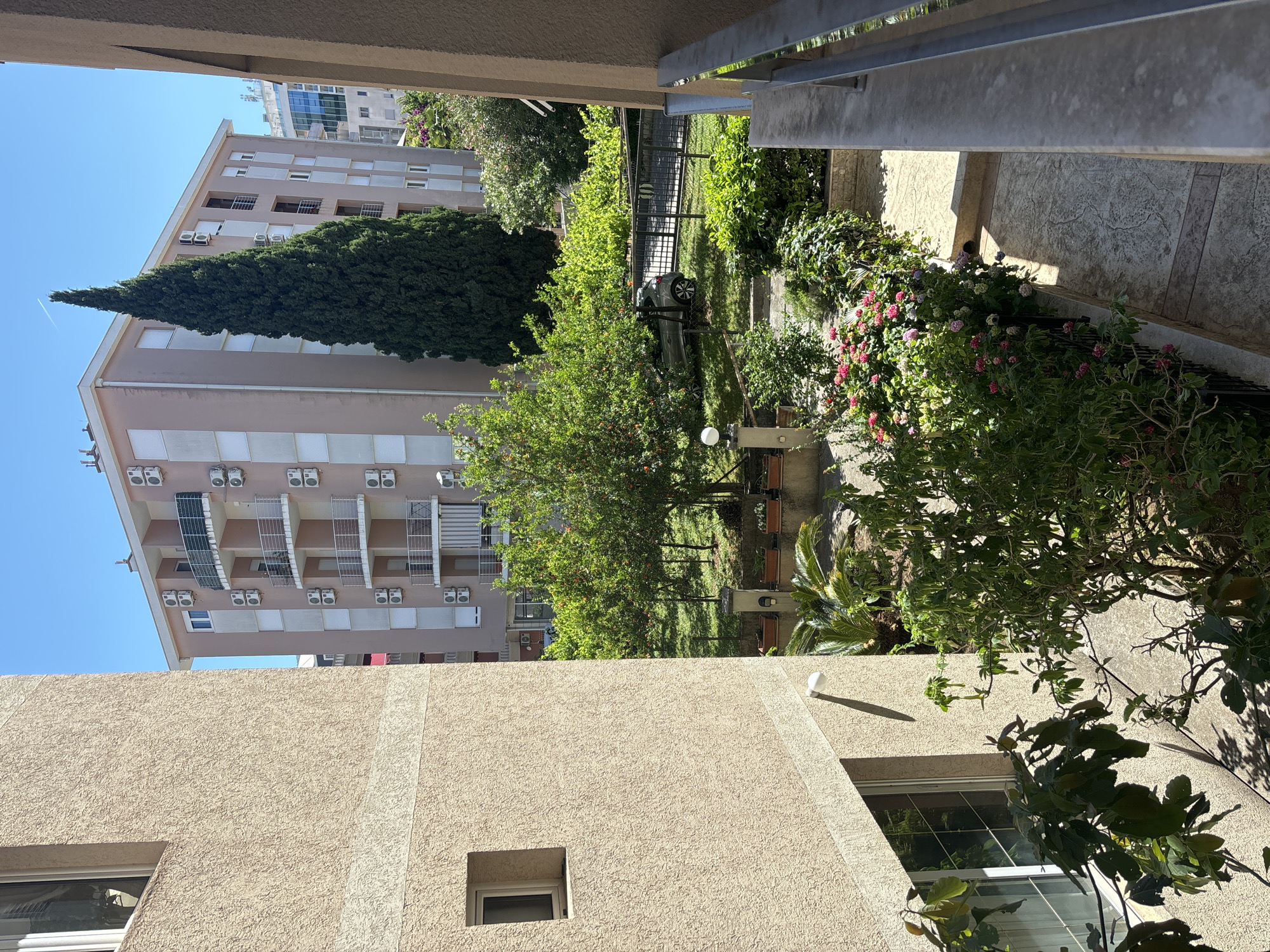 The photograph presents an urban landscape, likely taken from an elevated position in Budva, Montenegro. The central focus is a view between buildings, revealing a courtyard or garden area.
**Buildings:** Two buildings dominate the scene. One building has a beige exterior, with a small square window and a larger rectangular window visible on the right side. The second building is taller, with multiple stories and a lighter facade. Air conditioning units are attached to the walls beneath the balconies.
**Courtyard/Garden:** The view between the buildings exposes a green space. There are various trees, bushes, and some flowers. A dark-colored car is parked in the courtyard.
**Environment:** The scene is bathed in daylight, suggesting a sunny day. The sky is a clear blue. Shadows are visible, indicating the direction of the sunlight.
**Miscellaneous:** There is a metallic railing visible in the top-right of the image, suggesting the photograph was taken from a balcony or similar structure. A light fixture is attached to the building.