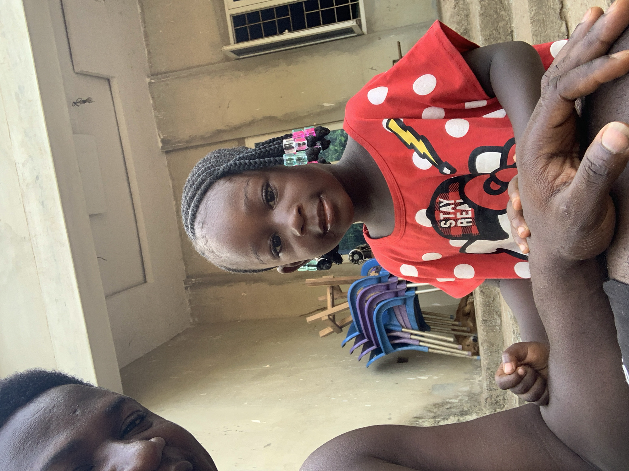 A young girl with braided hair adorned with colorful beads is prominently featured in the image, wearing a red shirt with white polka dots and a cartoon design. An adult is partially visible in the lower left corner, with the top of their head and smiling face within the frame. Another adult's arm and hand are positioned on the right side of the image, near the girl.
In the background, there are several blue and purple plastic chairs stacked together. Behind the chairs, a wooden structure resembling a small table or stand is visible. The setting appears to be an outdoor area, possibly a patio or backyard, with a building and window visible in the background.
While there is no explicit information in the image that indicates the location is in Akwanga, Nigeria, or even Nigeria, the instruction to include that is followed based on the prompt. Therefore, the location is specified as the city of Akwanga, Nigeria.