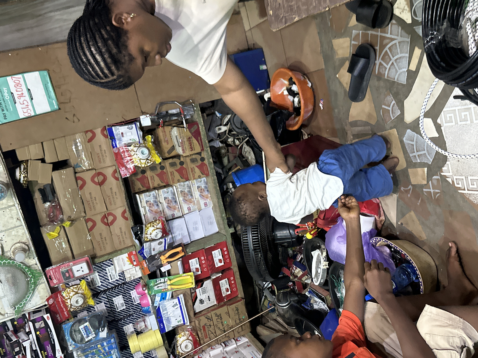 In Narayi, Nigeria, a crowded electronics and hardware store is captured, with shelves overflowing with merchandise. In the foreground, a young boy in an orange shirt and beige shorts sits on the floor, looking at a basket filled with various items. Beside him, a toddler in a white shirt and blue pants is being attended to by a woman with braided hair, who is wearing a white t-shirt and gold earrings. The woman appears to be holding the toddler's hand. The floor is covered with a mosaic of tiles and scattered debris, including black sandals and coiled wires. The background reveals more shelves packed with boxes, electrical components, and tools, suggesting a busy retail environment. A sign with a phone number and payment information is visible on a cardboard surface.