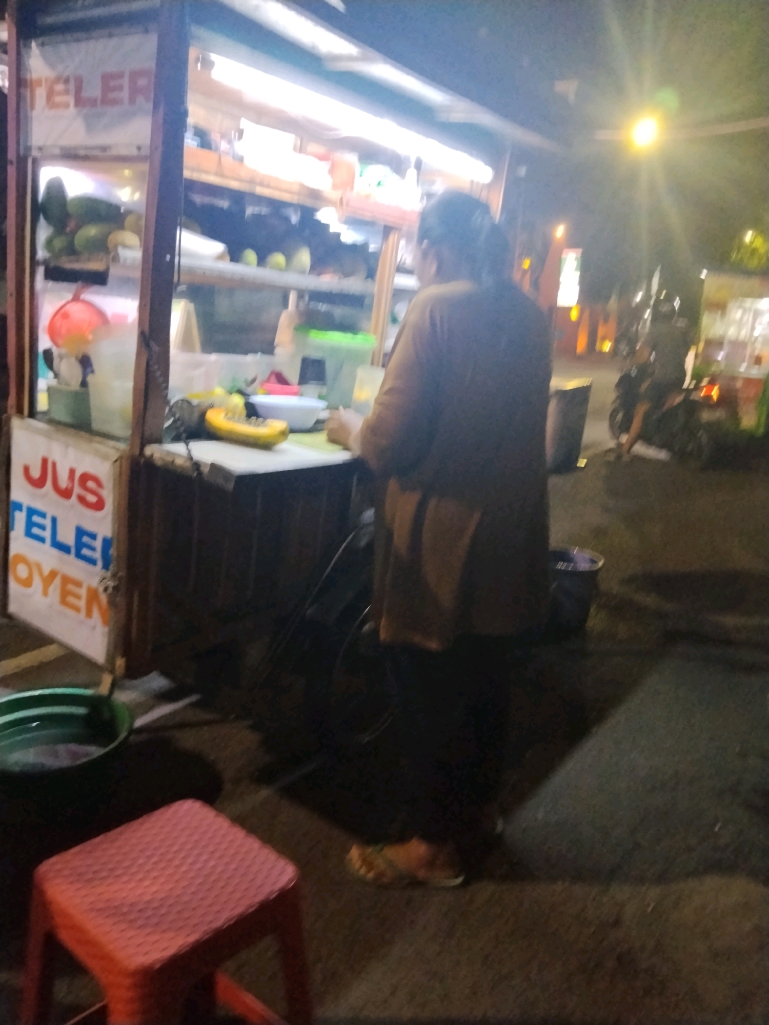In Mojokerto, Indonesia, at night, a food stall is seen from the side. The stall has a sign that reads "JUS TELE OYEN" and a display case with fruits. A person in a brown shirt is preparing food behind the stall. In the foreground, there is a red plastic stool and a green plastic basin. In the background, there are other stalls and a motorcycle. The lighting is dim, with a bright light source to the upper right of the image.