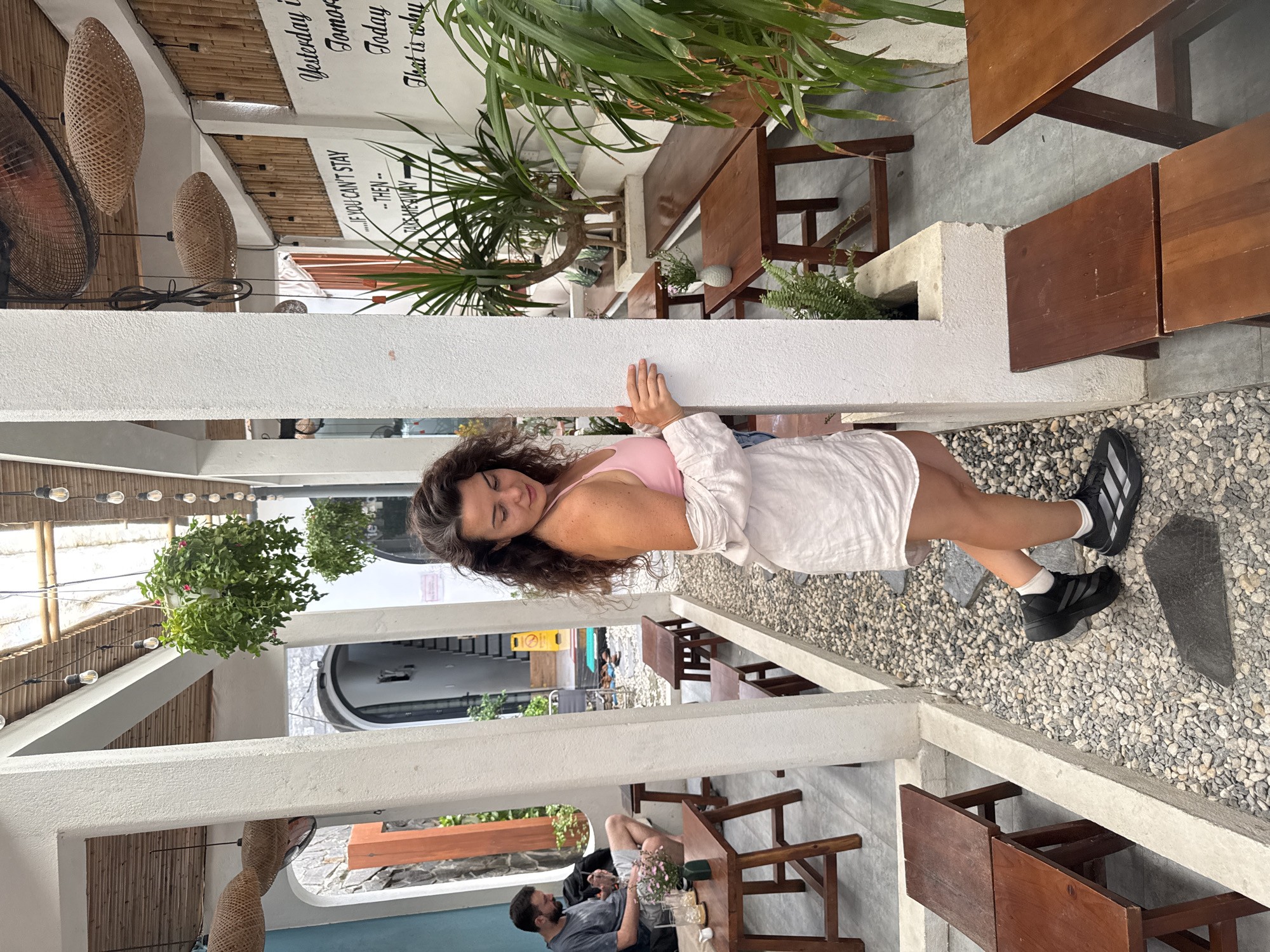 The image depicts an outdoor seating area of a cafe in Hoi An, Vietnam. In the foreground, a woman with dark, curly hair is standing and looking down. She is wearing a pink camisole, a light-colored unbuttoned shirt, denim shorts, and black athletic shoes with white socks. She is leaning against a white structural pillar. Behind her, a man with dark hair and a beard is seated at a wooden table on the left side of the frame, looking down at his phone. He is wearing a grey t-shirt and shorts. The cafe has a rustic design with thatched roof elements, bamboo accents, and exposed bulbs for lighting. Various potted plants are visible throughout the space, including hanging plants and larger indoor plants. Wooden tables and benches are arranged on a gravel and paved ground. In the background, a wall features text that appears to be motivational quotes. The overall atmosphere suggests a relaxed outdoor cafe setting.