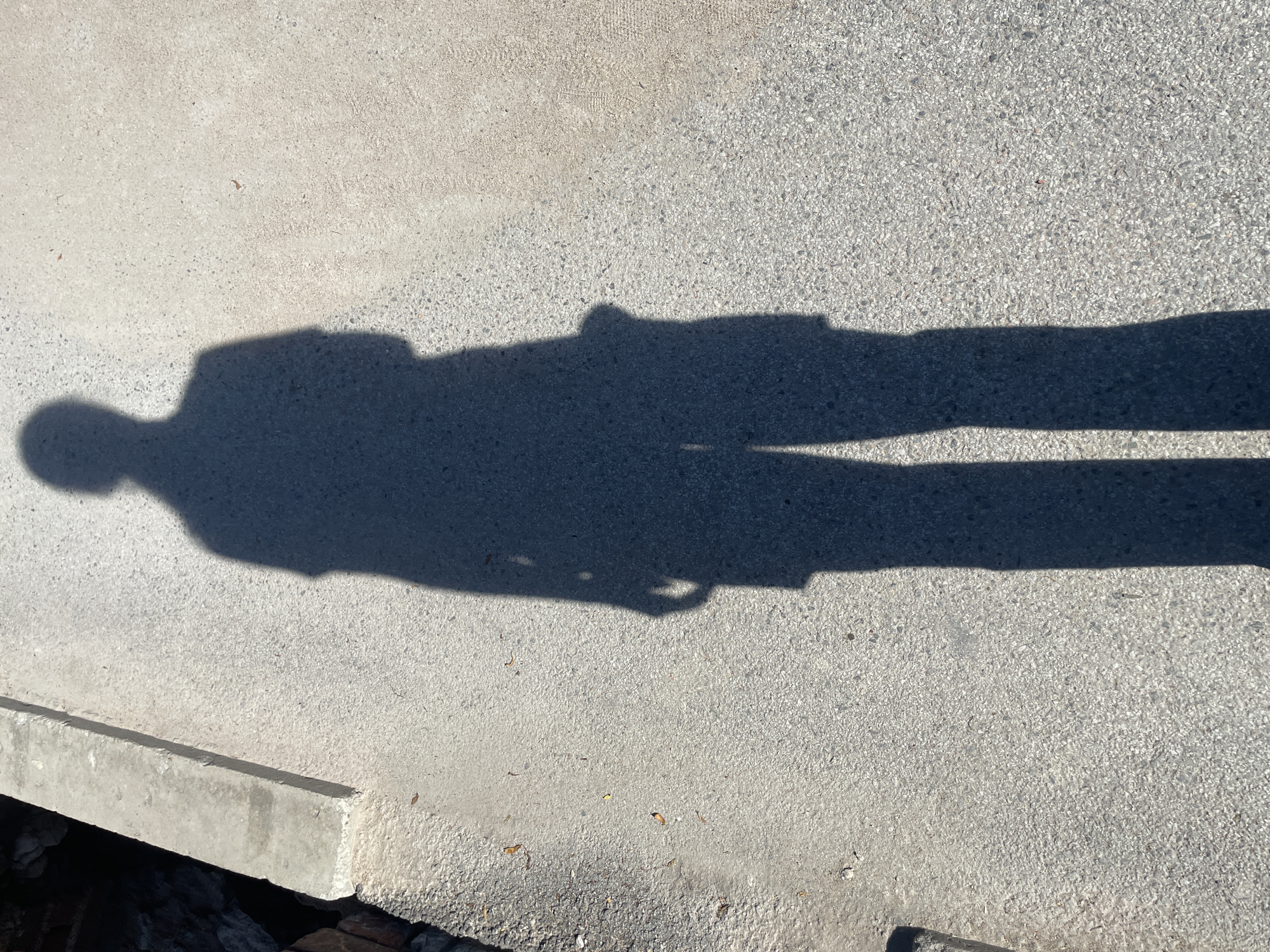 The image displays the long, distorted shadow of a person cast on a textured concrete surface. The shadow stretches horizontally across the frame, its head on the left and feet extending to the right. The figure appears to be wearing pants, and a distinct outline of a torso and head is visible. The concrete itself has a rough, granular texture, with some small pebbles or debris scattered across its surface. In the lower left corner, a concrete curb or edge is visible, with dark, rough material, possibly soil or rocks, beneath it. The bright sunlight suggests it is daytime, and the distinct shadow indicates clear weather. The overall scene is an outdoor setting, likely a sidewalk, path, or paved area. There is no visible text in the image.