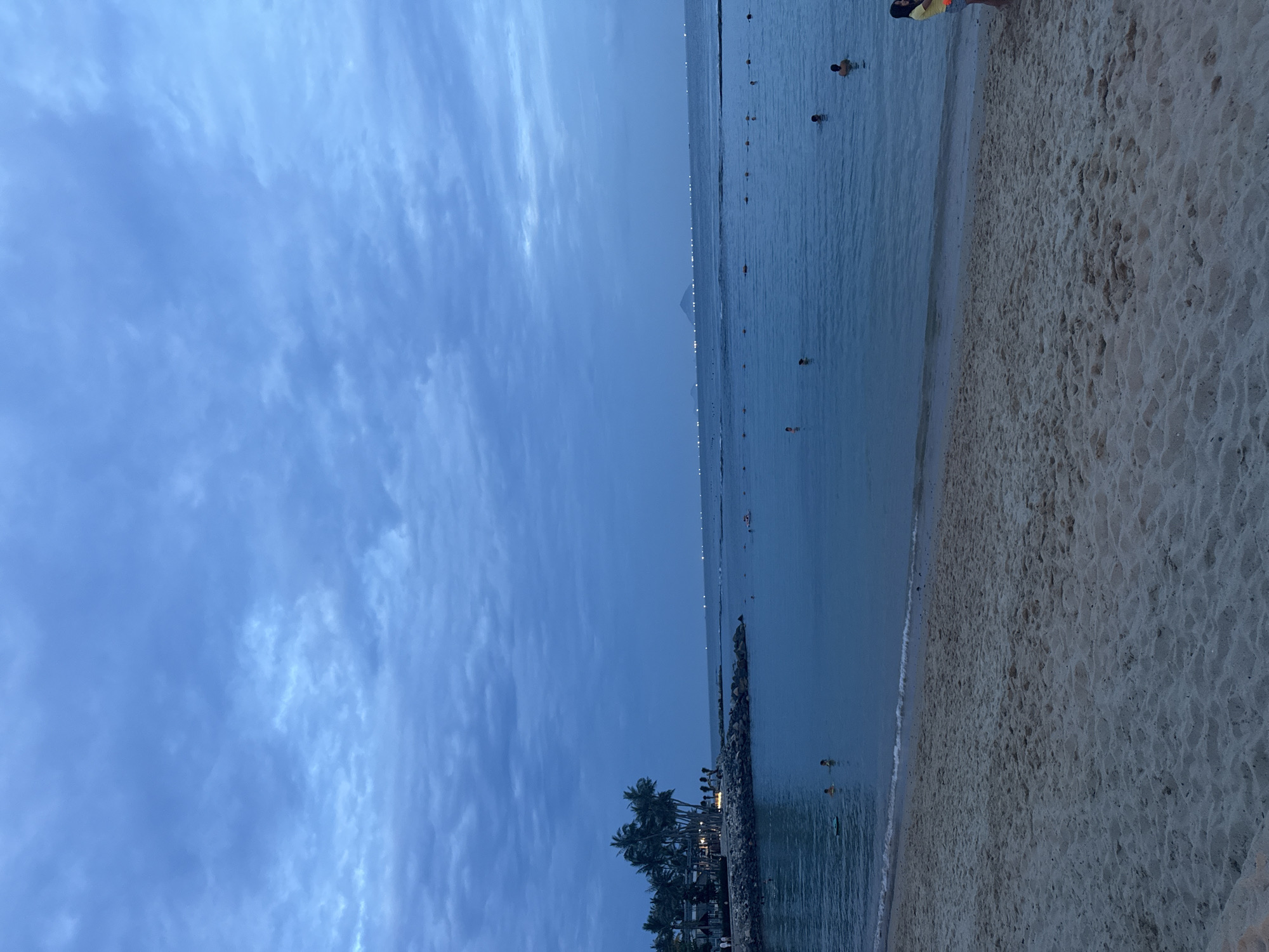 The image depicts a serene beach scene at dusk, with a body of water extending to the horizon under a cloudy sky. In the foreground, a sandy beach stretches across the bottom of the frame. The water is calm, with several individuals swimming and wading. To the left, a rocky embankment lined with palm trees and structures suggests a resort or populated area. Distant lights dot the horizon, possibly indicating a city or other coastal settlements. The overall mood is peaceful and tranquil.