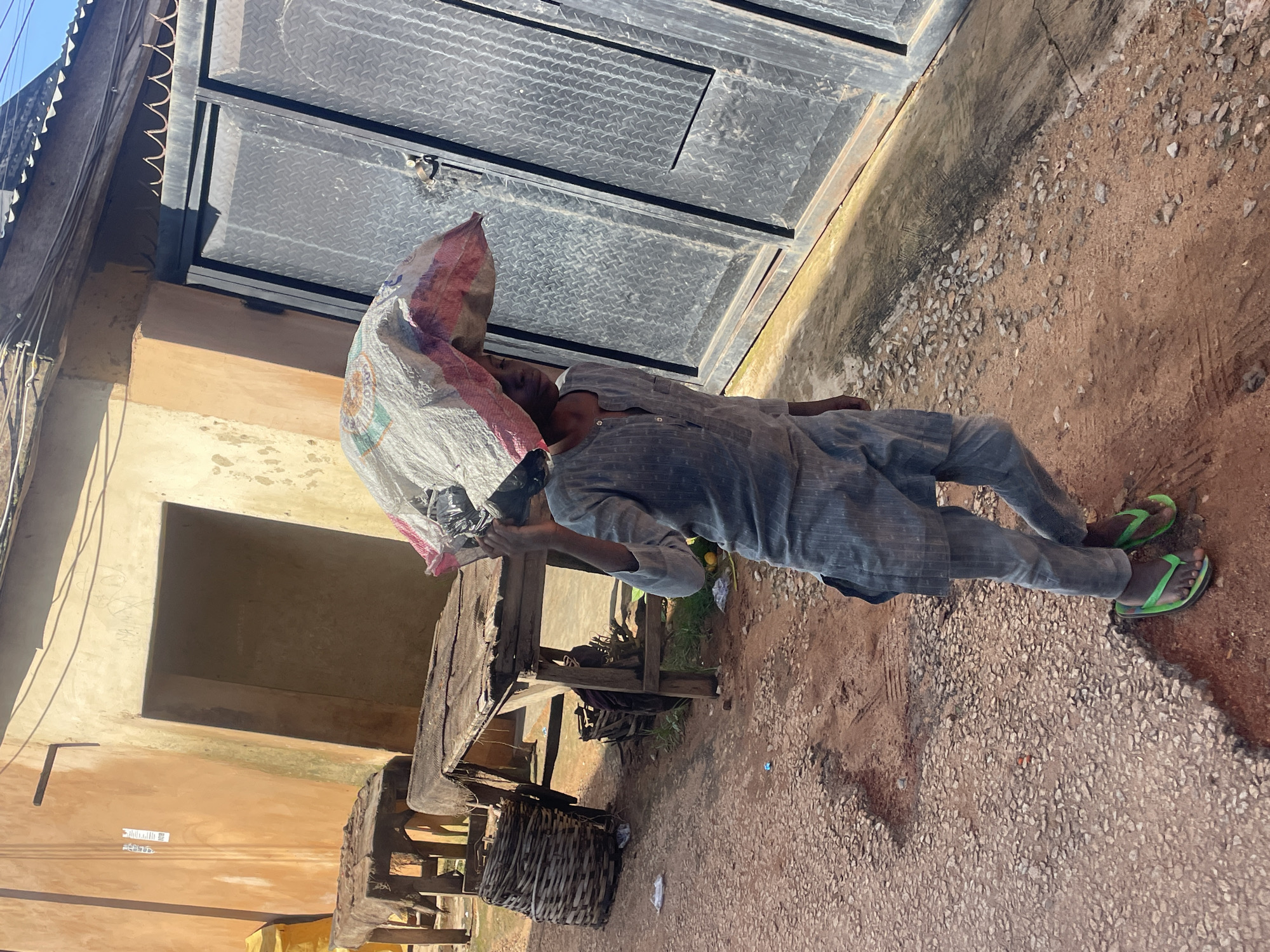 A person walks on a gravelly path in what appears to be an outdoor market area in Jos, Nigeria. The individual is wearing a grey, patterned outfit consisting of a tunic and trousers, along with green flip-flops. They are carrying a large, light-colored sack on their head, partially obscuring their face. Behind the person, wooden market stalls with worn coverings are visible, as well as woven baskets and a large metal gate. The buildings in the background are a light orange color, and the ground is a mix of red earth and small stones, with some scattered debris. The scene suggests a typical day in a Nigerian urban market.