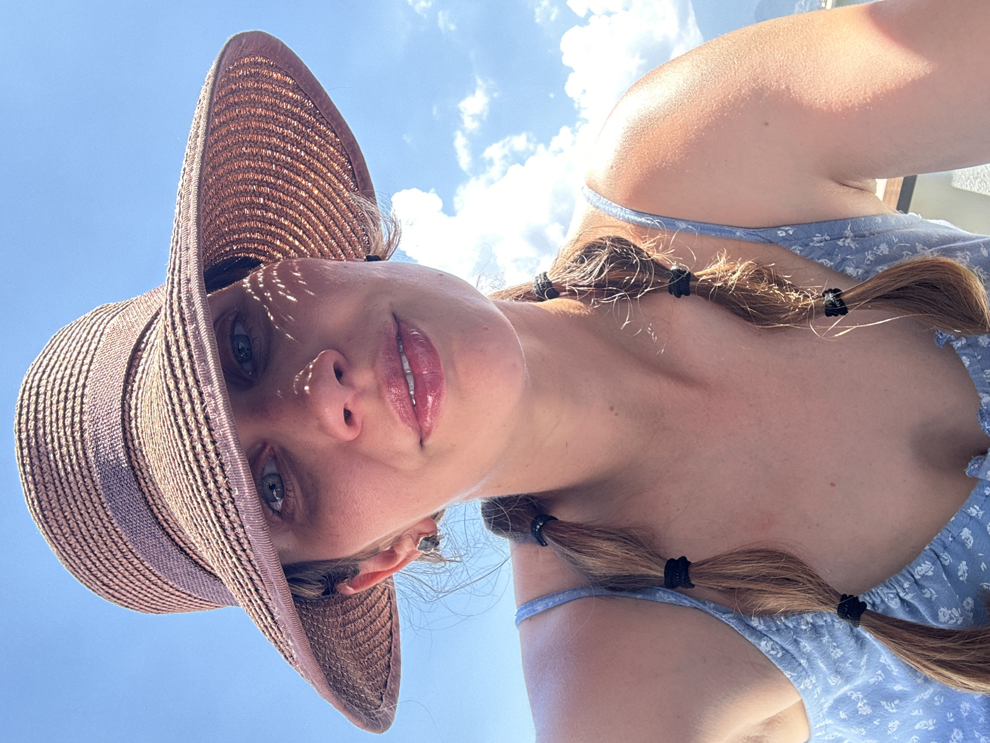 A young woman is captured in a close-up, low-angle shot against a bright blue sky with scattered white cumulus clouds. She wears a wide-brimmed, woven straw hat that creates intricate shadows from its texture across her forehead and face. Her light brown hair is styled in two pigtails, each secured with black elastic ties, resting on her shoulders and chest. She has fair skin, full, glossy pink lips, and light-colored eyes, looking directly towards the camera with a soft, slight smile. She is dressed in a light blue garment, possibly a dress or top, featuring a subtle white floral pattern and thin straps. The strong, natural sunlight highlights her features and arms, indicating a clear and sunny day. The perspective is from below, looking up, giving the image an open and expansive feel. The setting is Fethiye, Turkey.