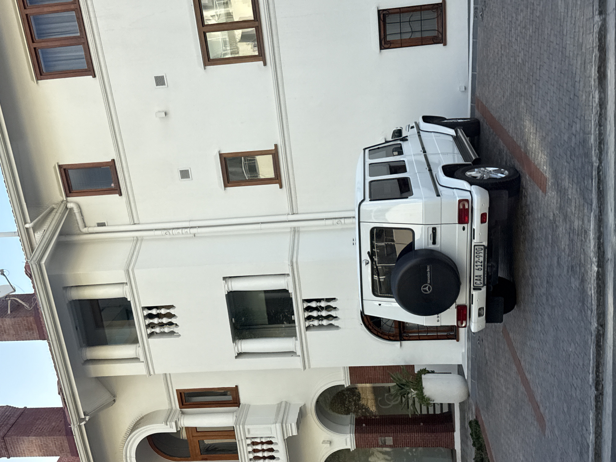 A white Mercedes G-Wagon is parked on a brick-paved street next to a white building. The building has multiple windows with brown frames. The license plate of the G-Wagon is visible and reads "CAA 612-990." Based on the architecture, the scene is likely located in the city of Cape Town, South Africa.