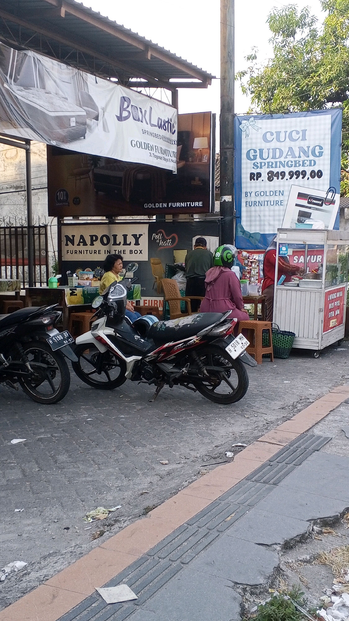 This image captures a street scene in Semarang, Indonesia, focusing on a sidewalk and street area in front of several businesses. In the foreground, two motorcycles are parked on a paved surface. The motorcycle on the right is white and black with colorful graphics, and its license plate reads "H 5018 MH". The motorcycle on the left is black, with a license plate reading "H 6198 RE".
Behind the motorcycles, a food cart is visible, with a sign displaying "SAYUR". A woman in a red shirt is attending to the cart. To the left of the food cart, a woman with dark hair is sitting. Behind them, there are various signs advertising furniture. One prominent sign reads "NAPOLLY FURNITURE IN THE BOX". Another sign, partially obscured, shows "GOLDEN FURNITURE". A blue banner with white text announces "CUCI GUDANG SPRINGBED RP. 849.999,00 BY GOLDEN FURNITURE". Another large banner to the left, draped, features an image of a mattress with the text "BoxLastic SPRINGBED".
The environment appears to be an outdoor commercial area, with a canopy overhead supported by poles. The ground is paved with asphalt and cobblestones, and some litter is scattered around. Trees and foliage are visible in the background, suggesting an urban setting. The overall impression is that of a busy street with ongoing commercial activity, likely during daytime given the lighting.