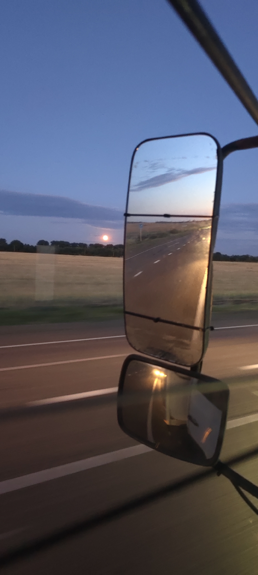 The image shows a view from inside a vehicle at dusk. The sky is a deep blue with some wispy clouds and a bright moon visible in the distance. The scene is reflected in a large side mirror of the vehicle, which shows a road stretching into the distance, illuminated by the headlights of the vehicle. In the reflection, fields of dry grass and a line of trees can be seen along the side of the road. A second, smaller mirror is visible below the large one, also reflecting the road and vehicle lights. The overall impression is one of a quiet journey through a rural landscape as night falls.