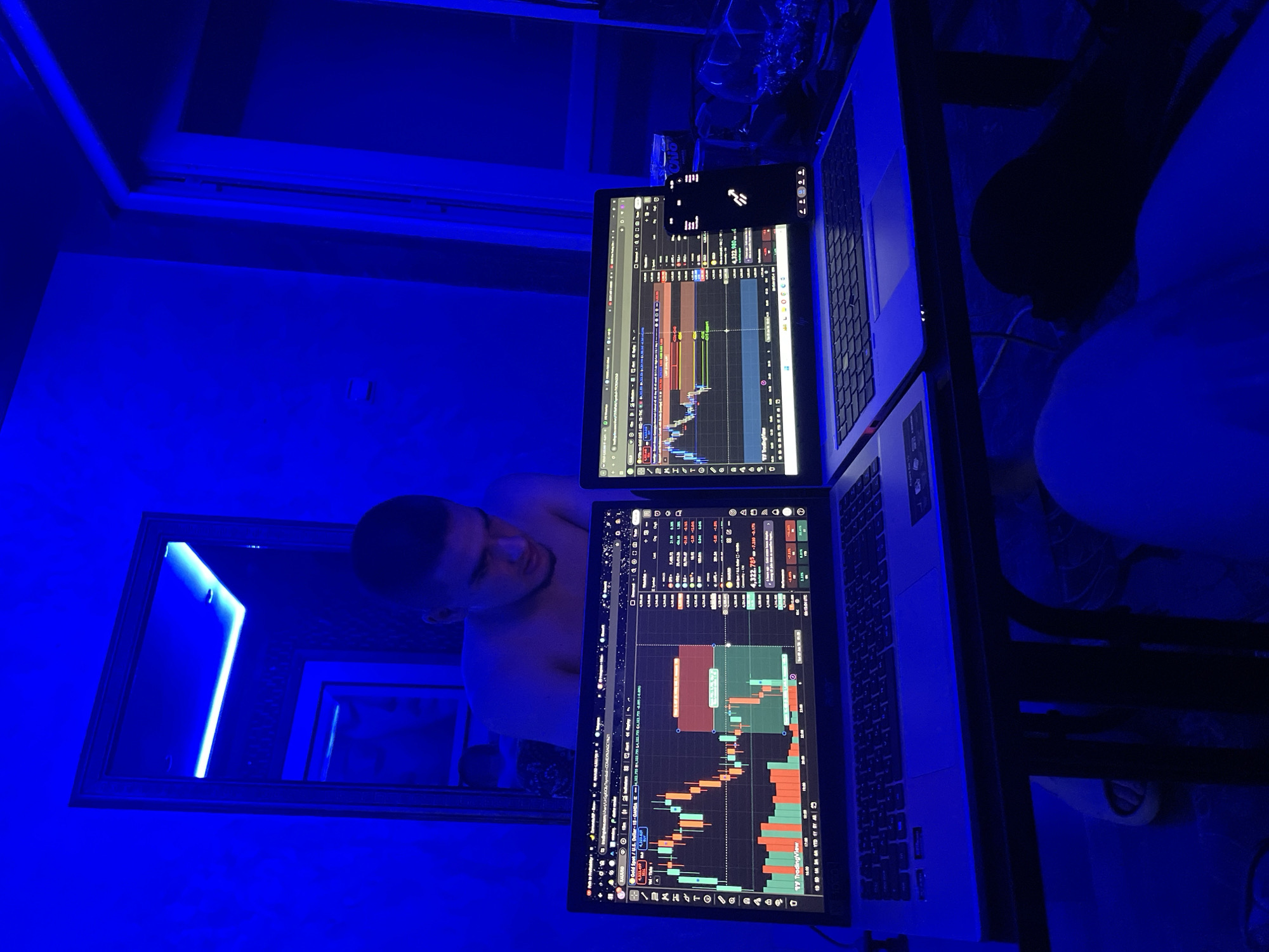 The image shows two laptops open on a dark table, displaying stock market charts. A young man is looking down at the screens, appearing to be engaged in trading. The room is dimly lit with blue LED lighting, creating a dramatic atmosphere. The overall scene suggests a home office or private workspace. The provided location of Asenovgrad, Bulgaria, cannot be verified from the visual information alone.