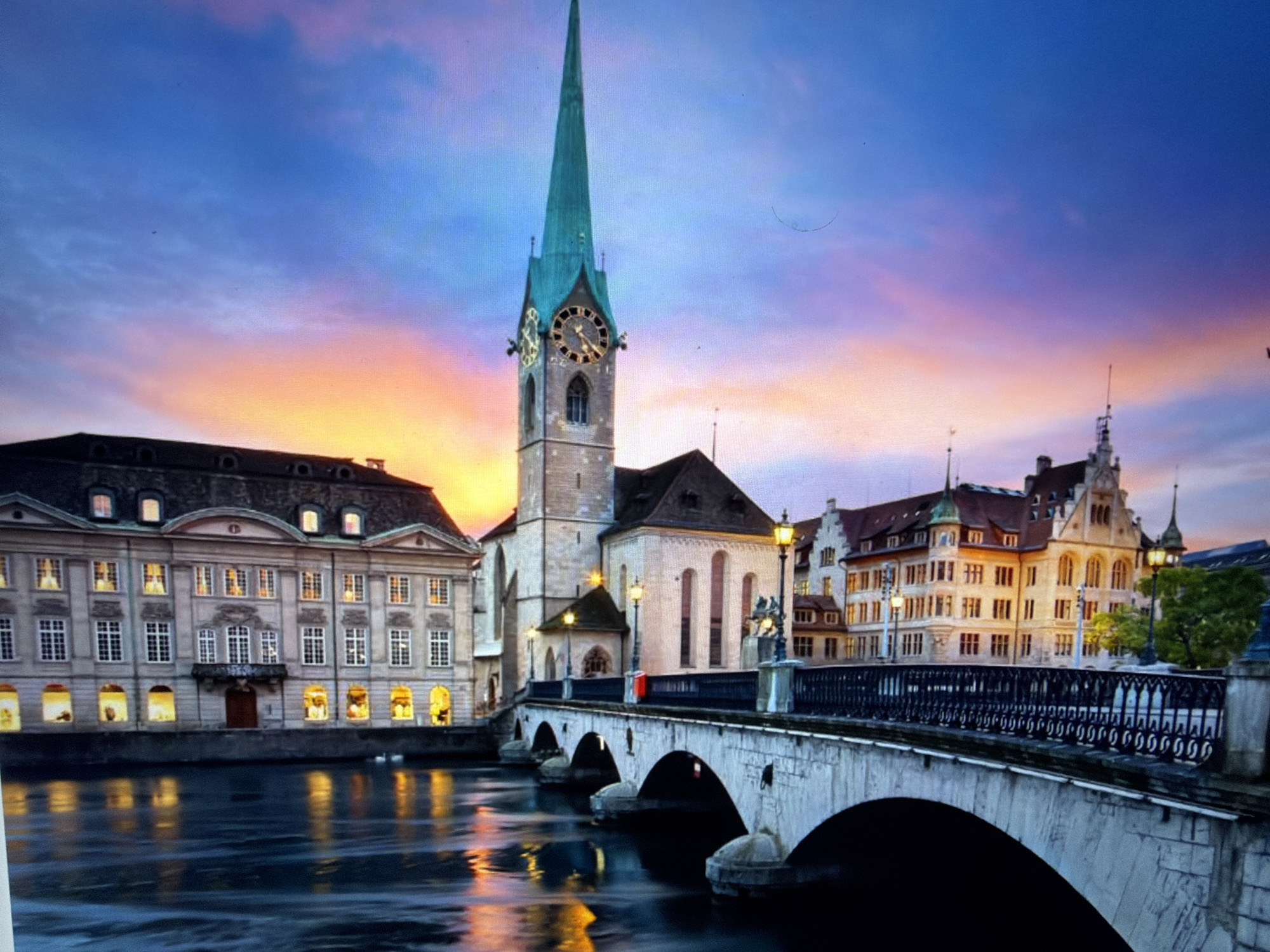 The image depicts a picturesque canal scene at twilight. A prominent stone bridge with multiple arches spans the water, reflecting the warm glow of streetlights and illuminated buildings. On the left, a grand stone building with numerous windows, some brightly lit, lines the canal. To the right of the bridge, a tall church tower with a green spire dominates the skyline, featuring a clock face and Gothic architectural elements. Behind the bridge, a row of historic buildings with ornate facades and many windows adds to the charming urban landscape. The sky displays a gradient of colors, transitioning from deep blue at the top to vibrant orange and pink hues near the horizon, indicating sunset or sunrise. The water is dark and slightly rippled, carrying reflections of the surrounding lights. The scene is devoid of any visible people or animals. The location is identified as Falenty Nowe, Poland.