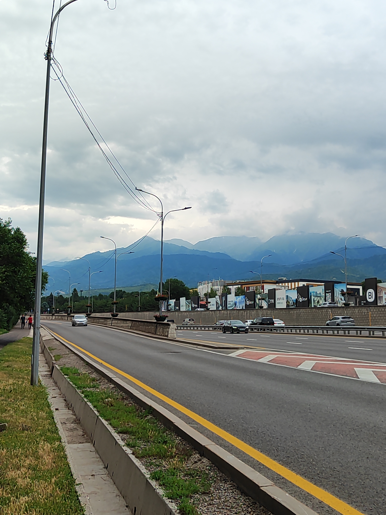 A wide-angle, eye-level shot captures a cloudy day in Almaty, Kazakhstan, featuring a multi-lane highway with cars driving. In the foreground, a grassy area with a concrete curb lines the left side of the road. A prominent lamppost stands tall on the left. The highway itself is paved, with yellow lines marking lanes. Several streetlights are visible along the road. In the middle ground, a low concrete barrier separates the highway from a pedestrian walkway on the far left, where two individuals are walking. Beyond the highway, buildings with large advertisements are visible. The background is dominated by majestic, hazy blue mountains, with some appearing snow-capped, suggesting the presence of the Tian Shan mountain range. The overall atmosphere is overcast and slightly muted due to the cloudy sky.
