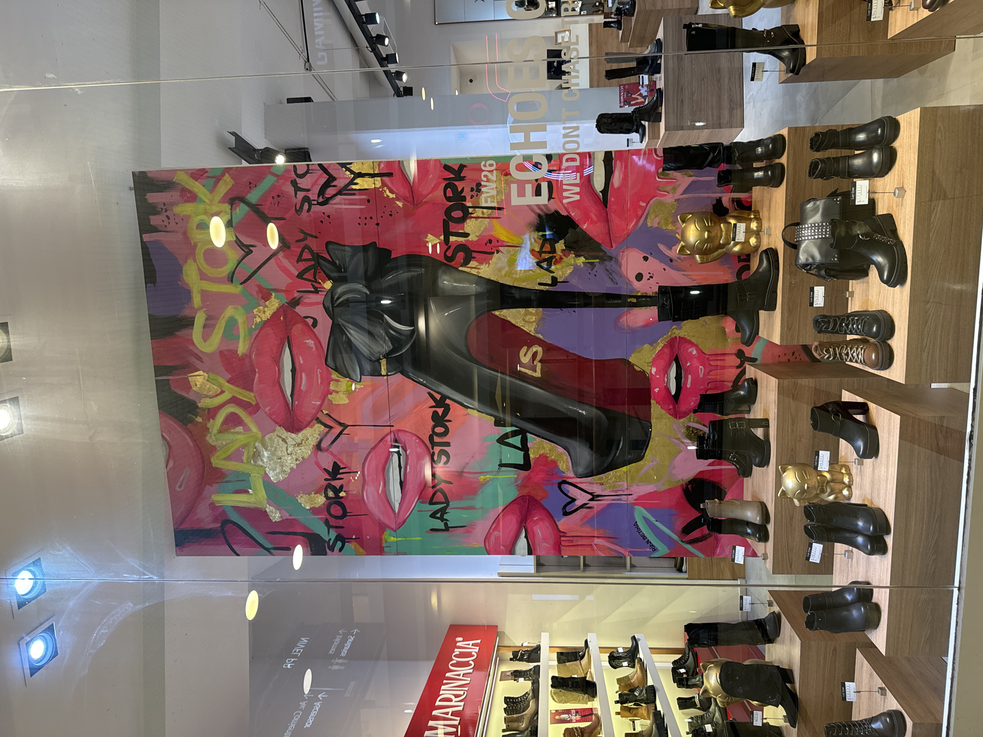 The image depicts a storefront display in Buenos Aires, Argentina. The store, identified by the "MARINACCIA" sign, showcases a variety of women's footwear, primarily boots, arranged on shelves. A large, colorful advertisement is on the wall, featuring an illustration of a woman's shoe, along with text that reads "LADY STORK," "ECHOS," and "WE DON'T CHASE." The advertisement includes artistic elements such as lips, graffiti-style text, and splashes of gold leaf. Each pair of shoes is labeled with a price. A golden cat figurine is displayed next to the boots on the display. The storefront is visible through a window, with reflections of the surrounding environment visible in the glass.