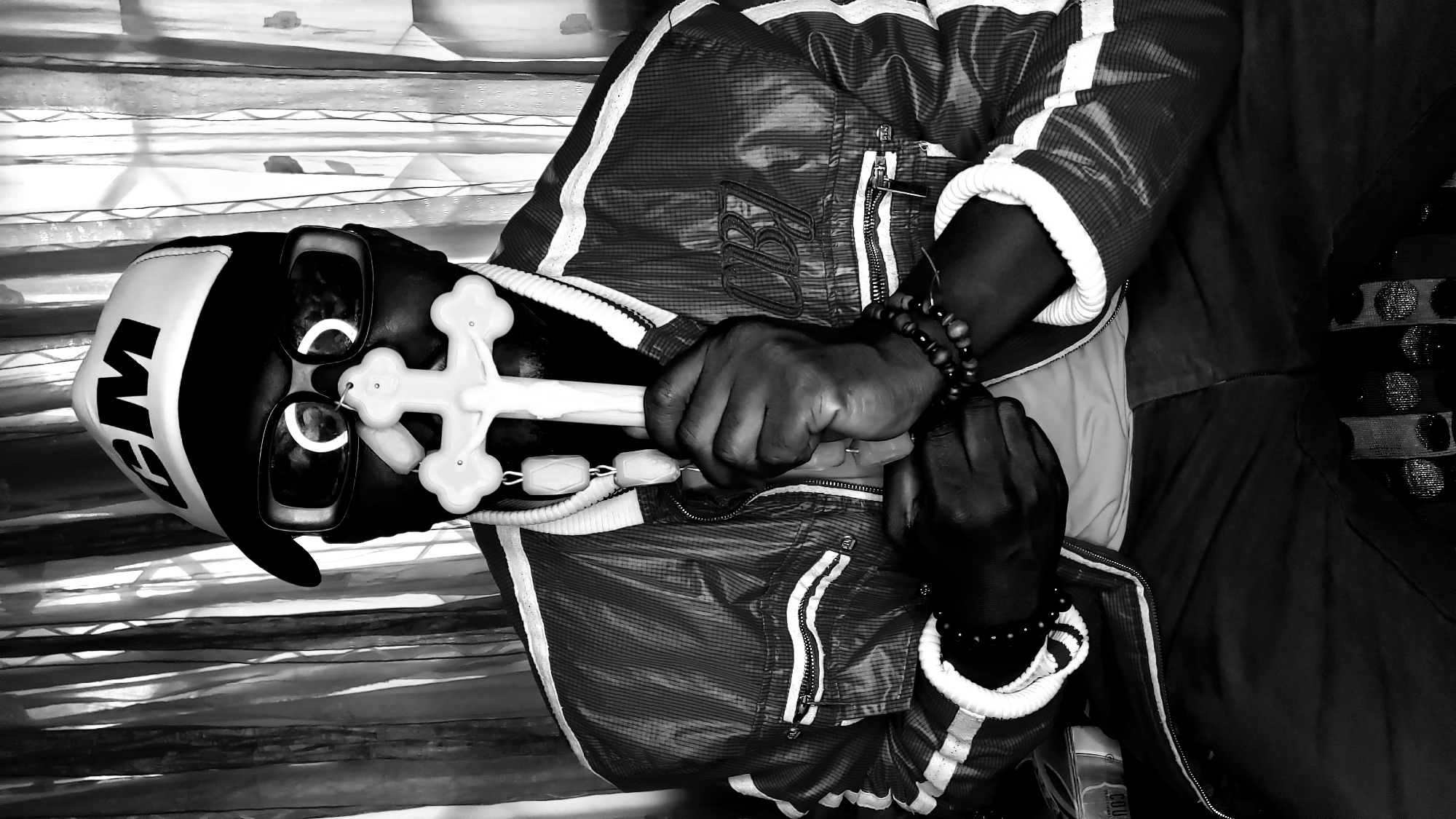 A black and white, medium shot photograph captures a person sitting indoors, holding a white cross up to their face. The person is wearing a black baseball cap with "CM" in white letters, large dark sunglasses with white circular reflections in the lenses, and a patterned jacket with white stripes on the sleeves and collar. The person's hands are clasped around the cross, and they are also wearing beaded bracelets on their wrists. A beaded necklace hangs from the cross. The background consists of vertical drapes or curtains. The scene is lit with dramatic lighting that casts strong shadows. The photograph appears to have been taken in Sabon Tasha, Nigeria.